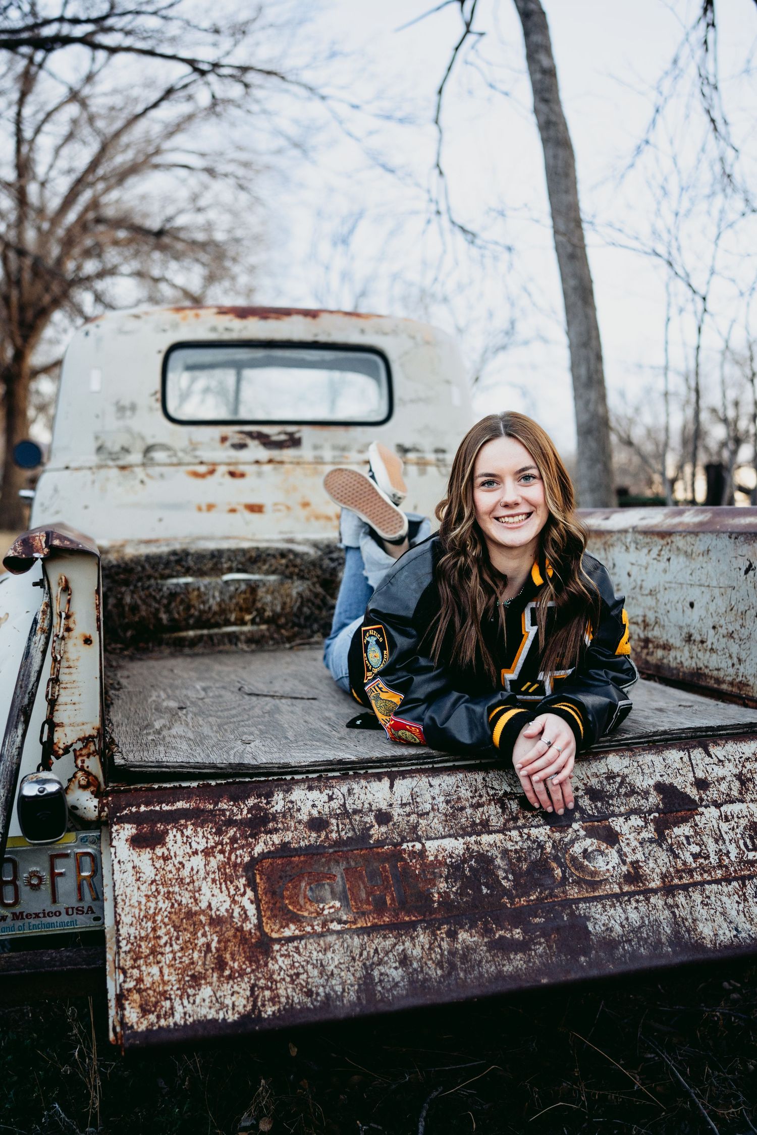 Happy high school senior girl posing in antique rusted pickup truck bed in black varsity jacket.