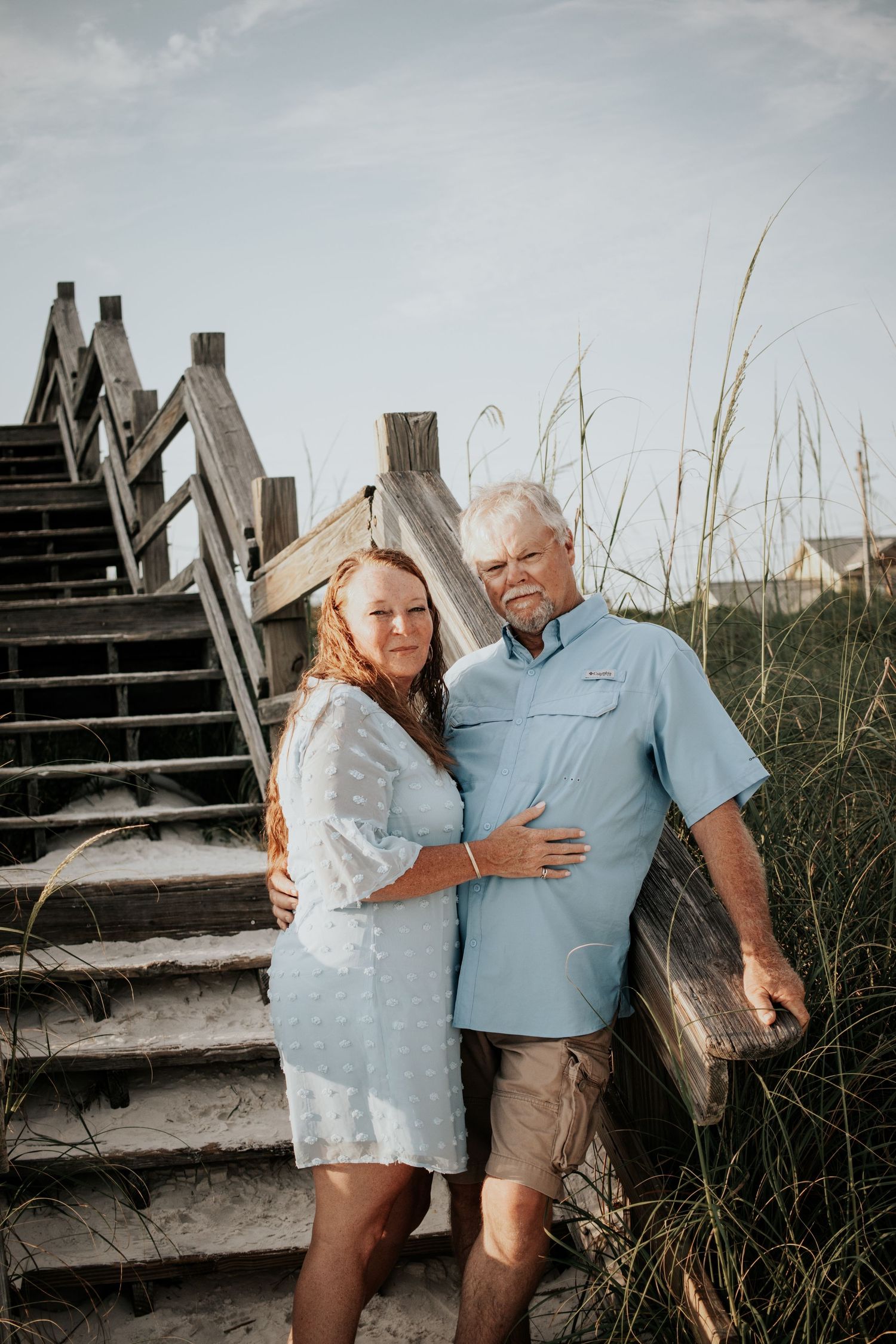 Mature couple posing together by wooden beach stairs among sea grass, woman in white dress, man in blue shirt.