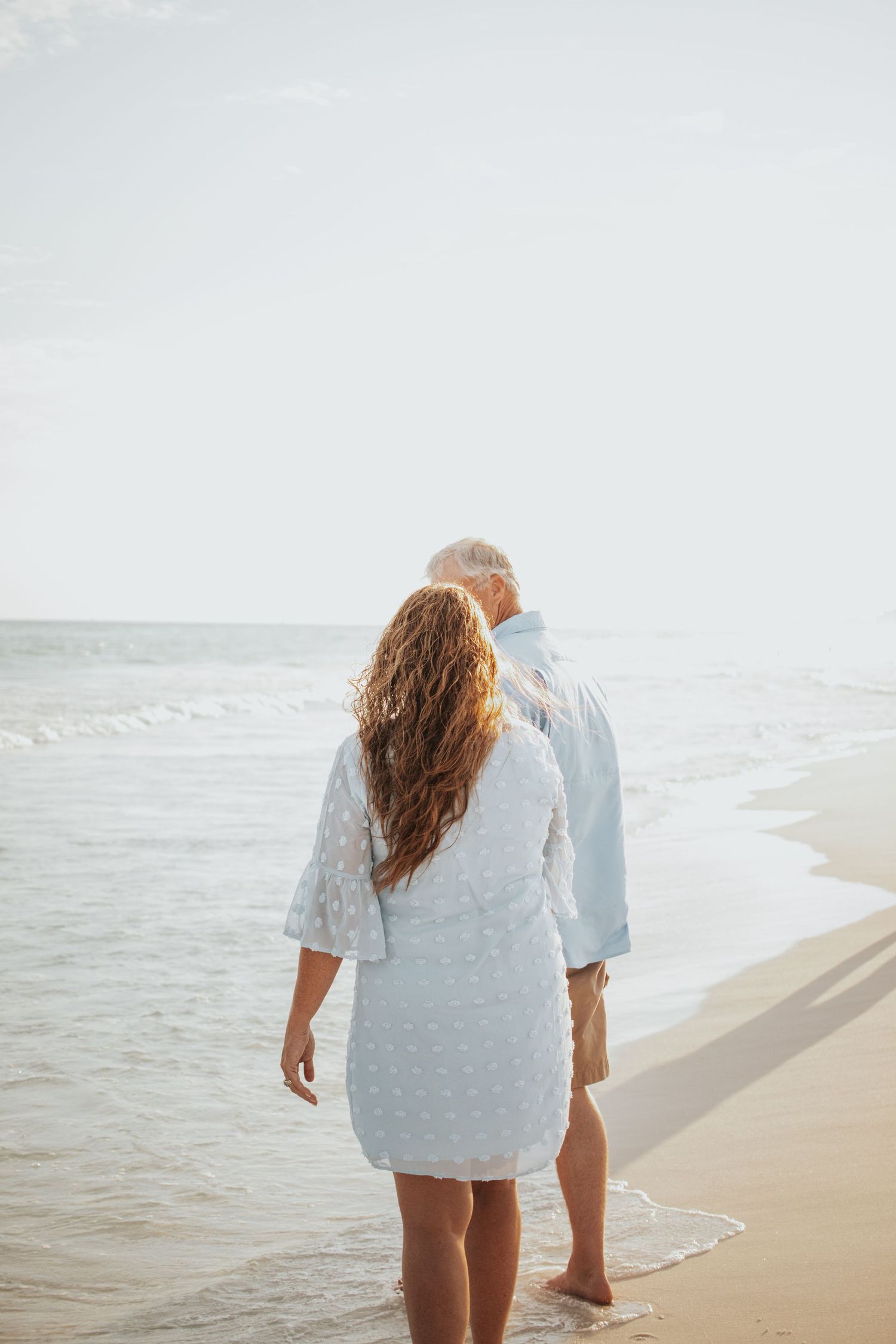 Mature couple walking barefoot along the shoreline at sunset, viewed from behind in matching blue outfits.
