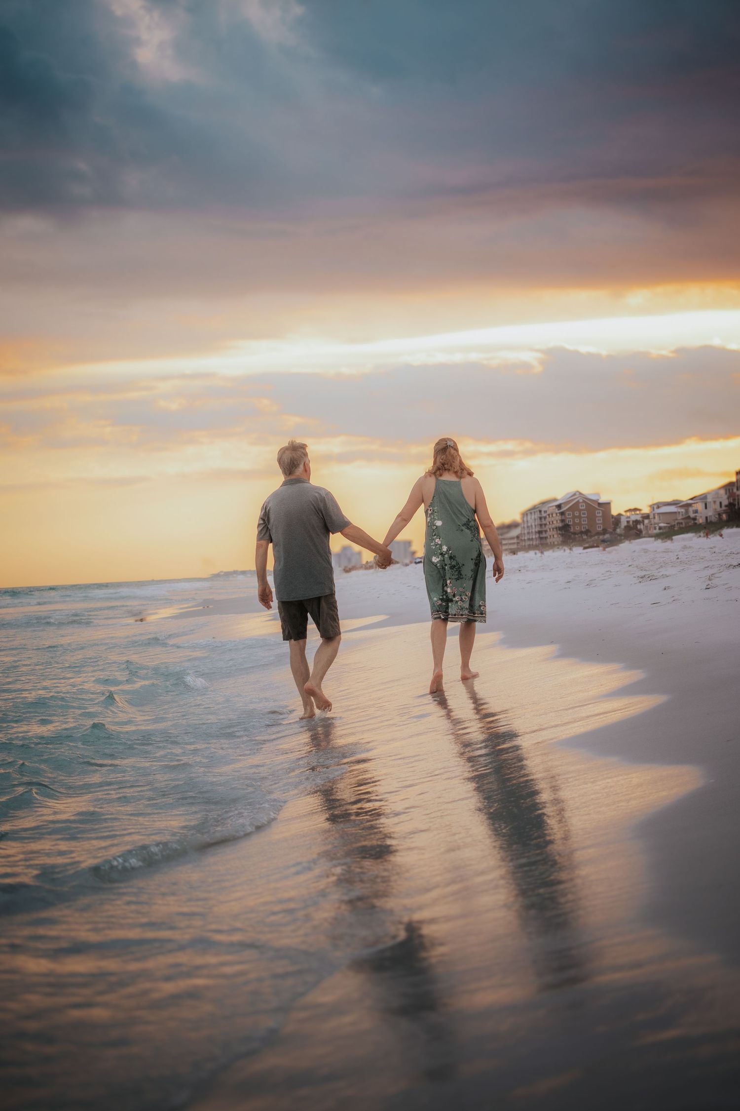 Couple walking hand-in-hand along a sandy beach at a colorful sunset with waves nearby.