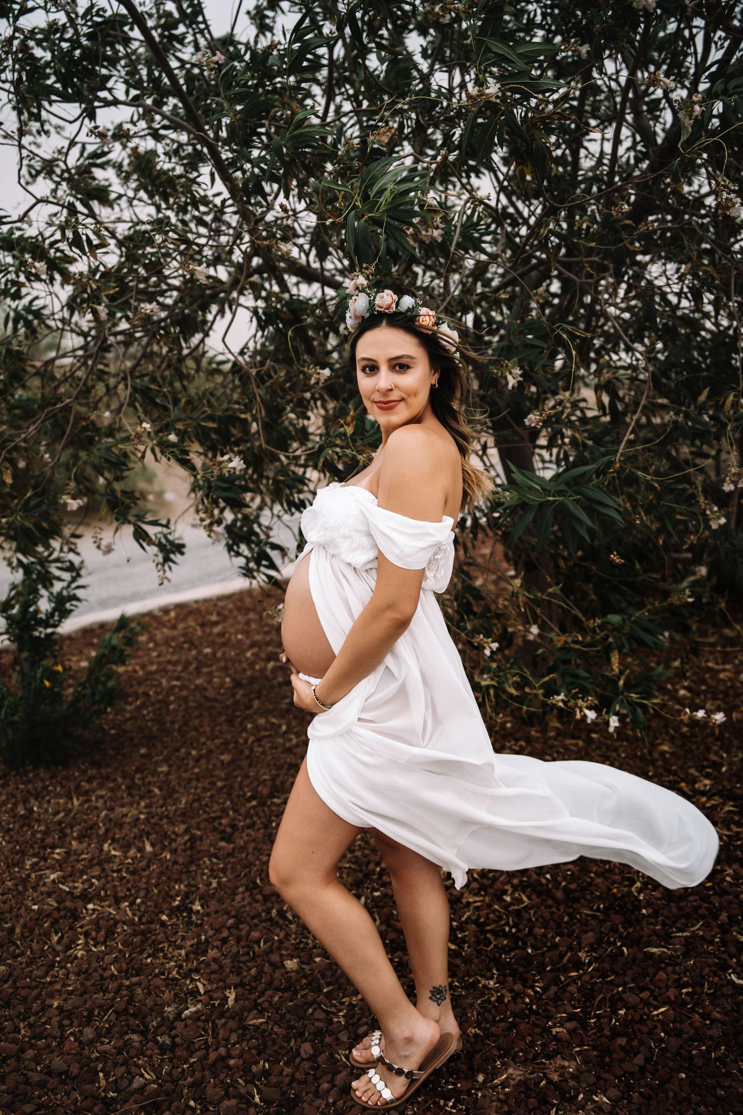 Pregnant woman in billowing white maternity dress and floral crown stands smiling before dense green foliage.