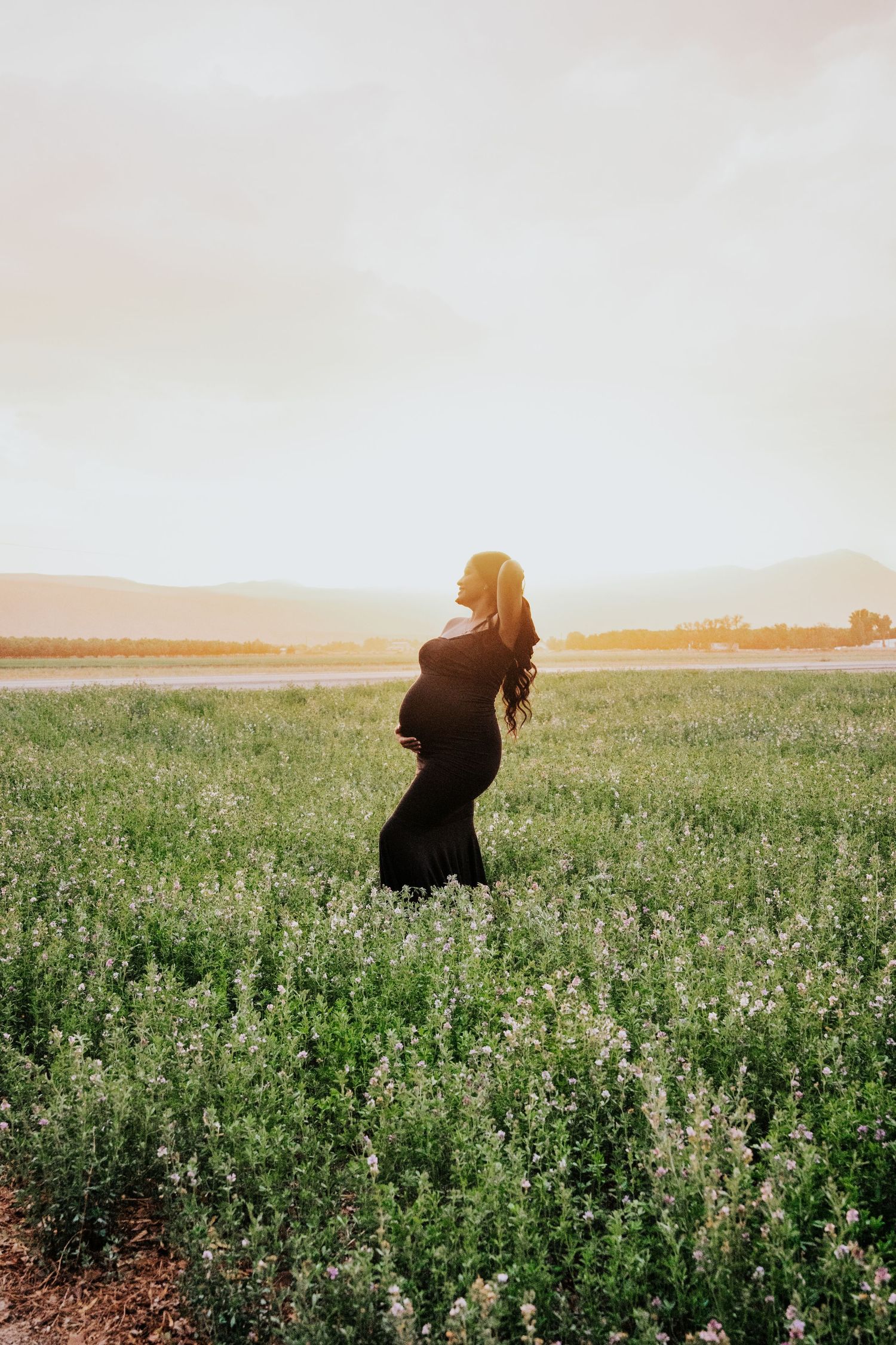 Pregnant woman in black gown standing in green flower field with mountains at sunset.