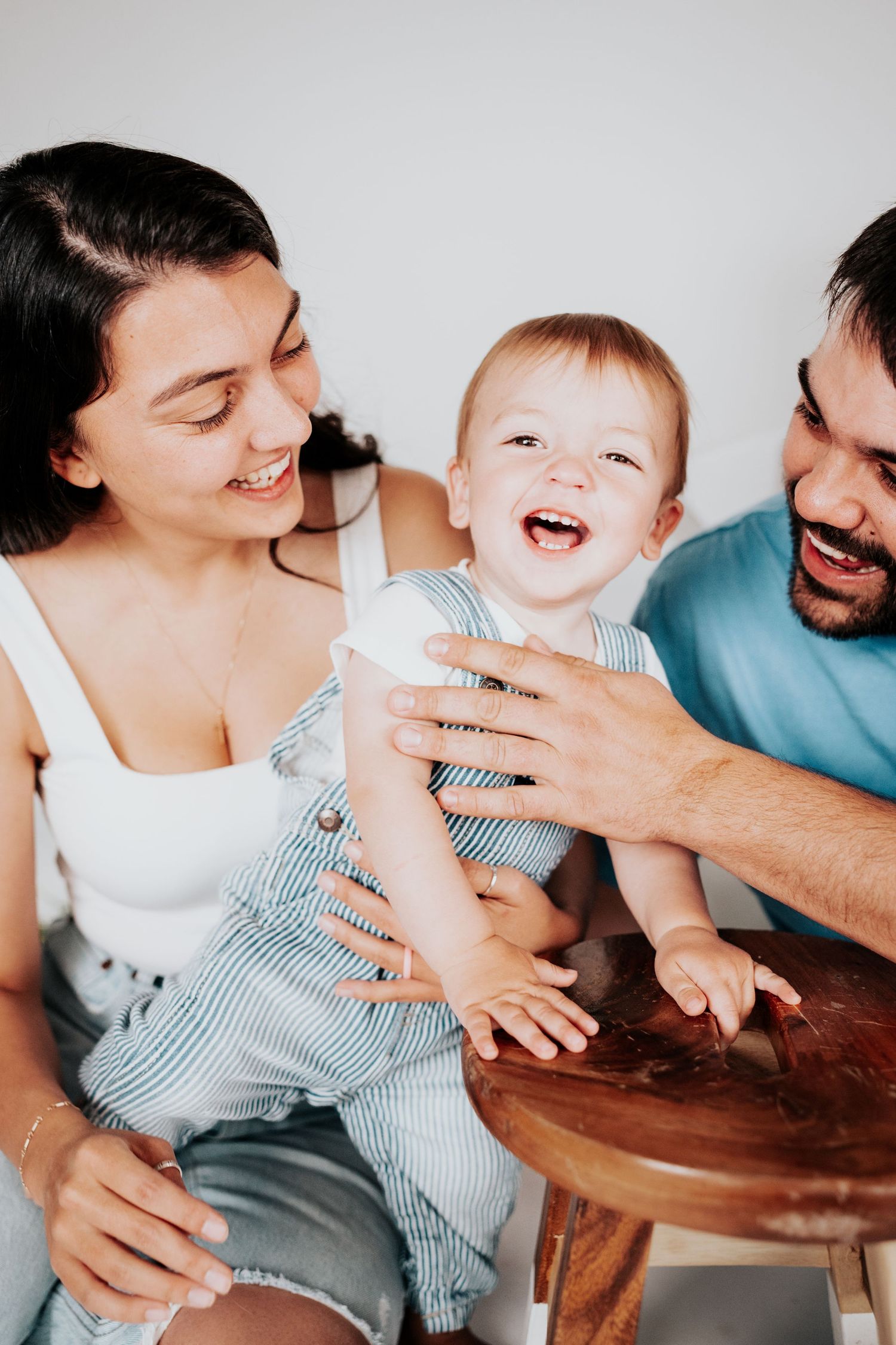 Joyful family moment as laughing baby in striped overalls delights parents during indoor photo session.