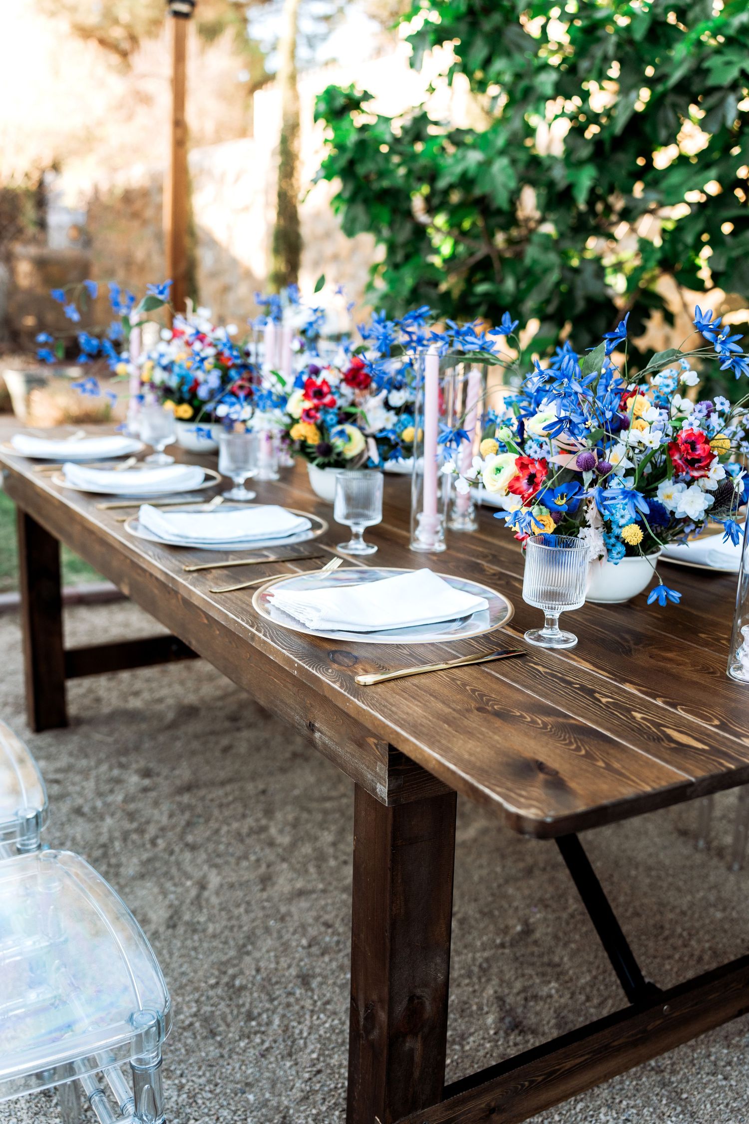Elegant outdoor wedding reception table with blue wildflower centerpieces, white plates, and gold cutlery on rustic wood.