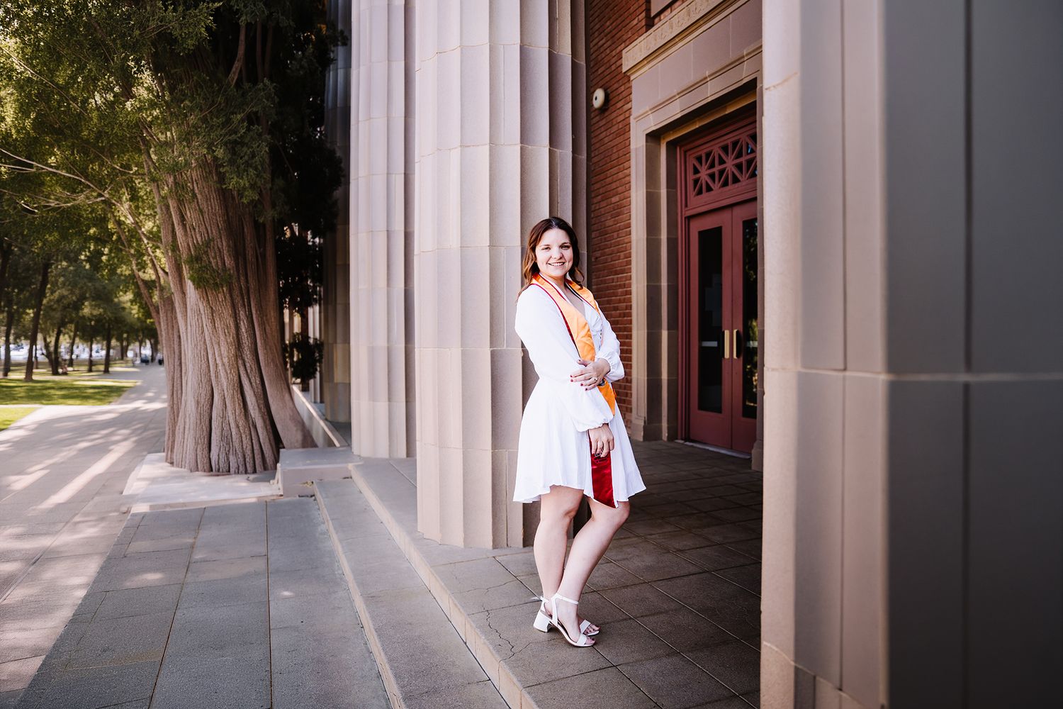 Young woman in white graduation dress with colorful sash poses confidently beside brick building columns on sunny campus.