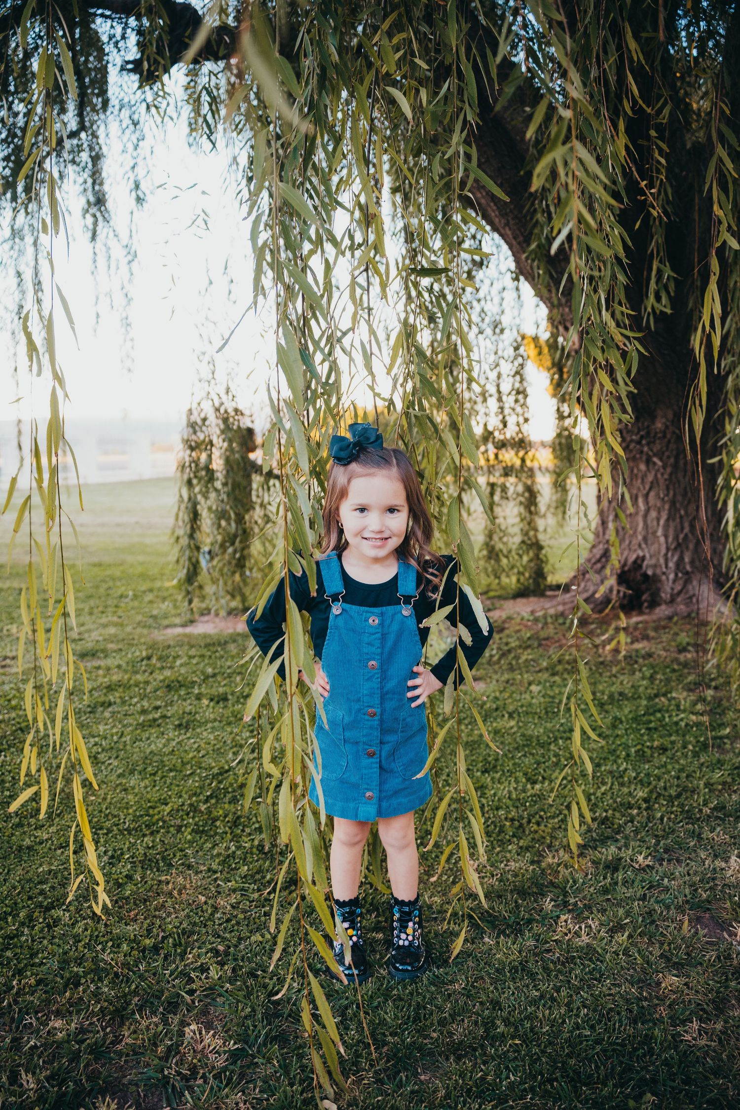 Smiling little girl in blue denim overalls standing under a weeping willow tree in a grassy park.