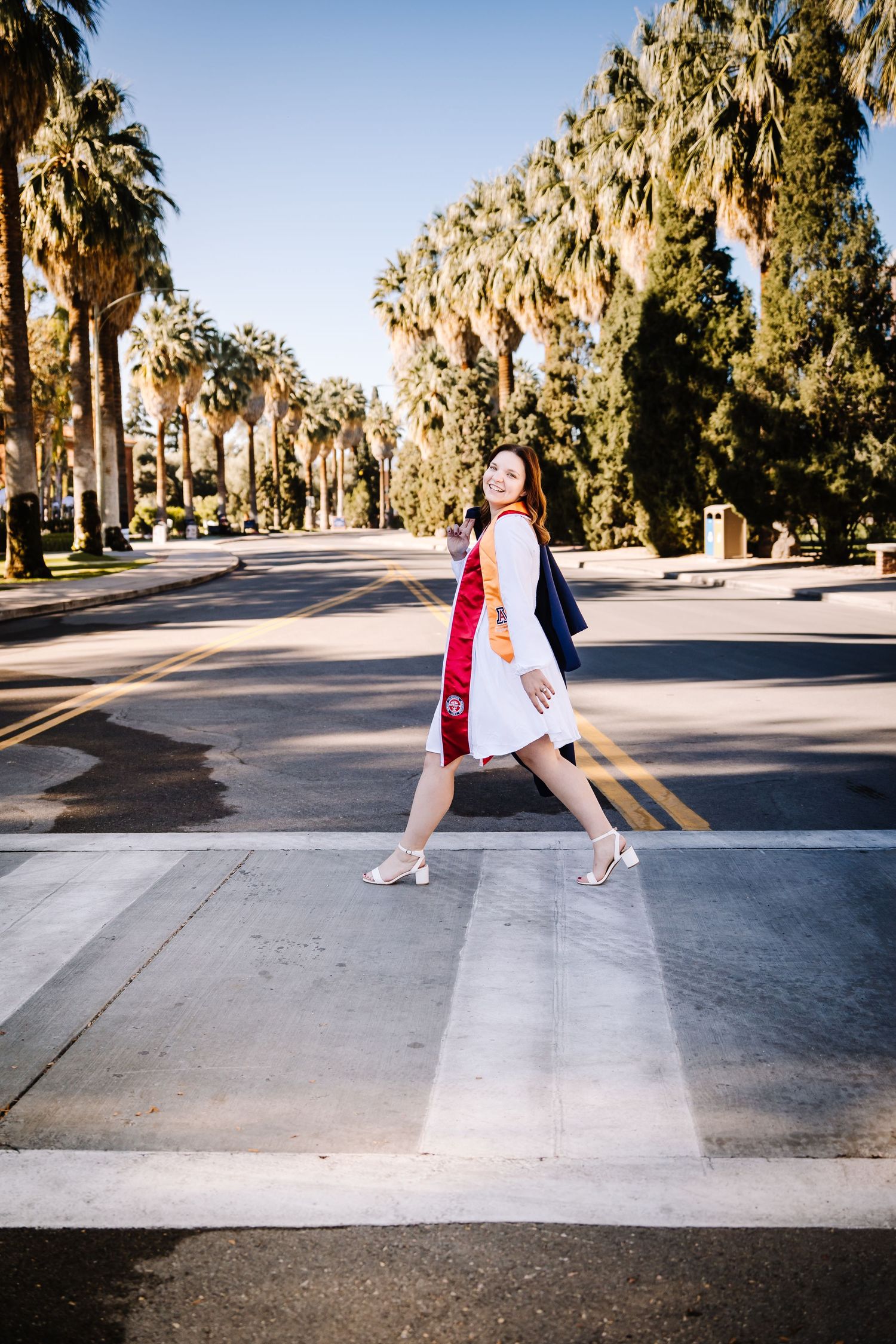 Female graduate in white dress and regalia crosses palm tree-lined street at university.