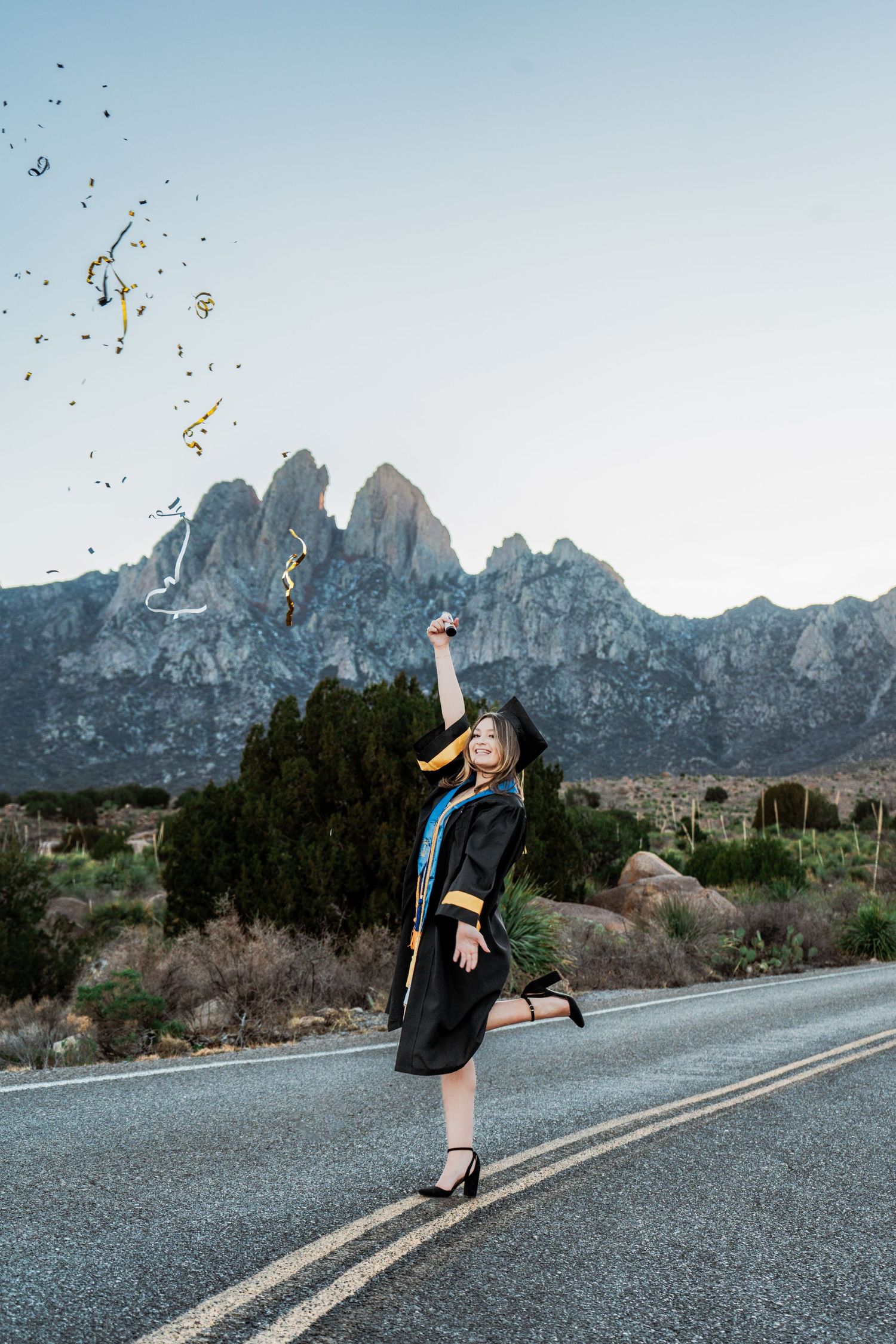 Graduate in cap and gown celebrating on a mountain road by tossing confetti in the air.