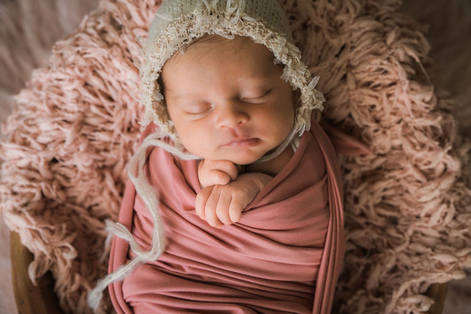 Peaceful newborn baby girl wrapped in dusty rose swaddle with delicate lace bonnet sleeping in basket.