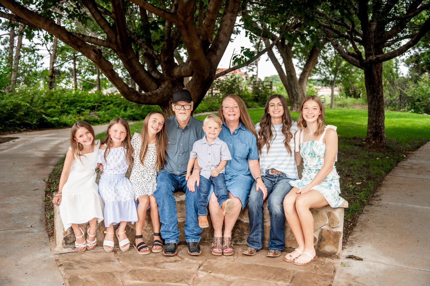 Family of nine posing on stone bench under large shady trees in park setting.