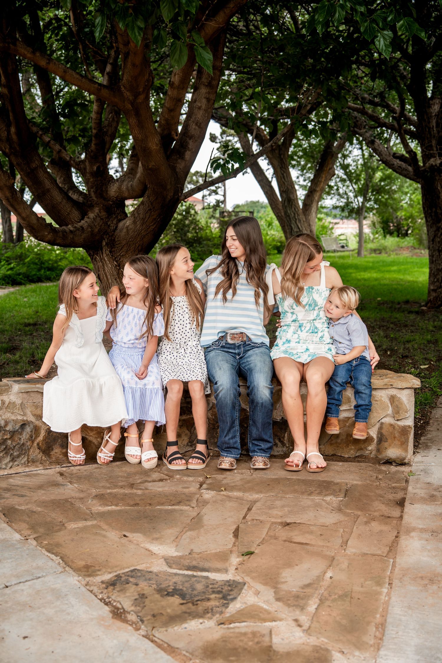 A woman sits on a stone bench with five young children under shady trees in a park setting.