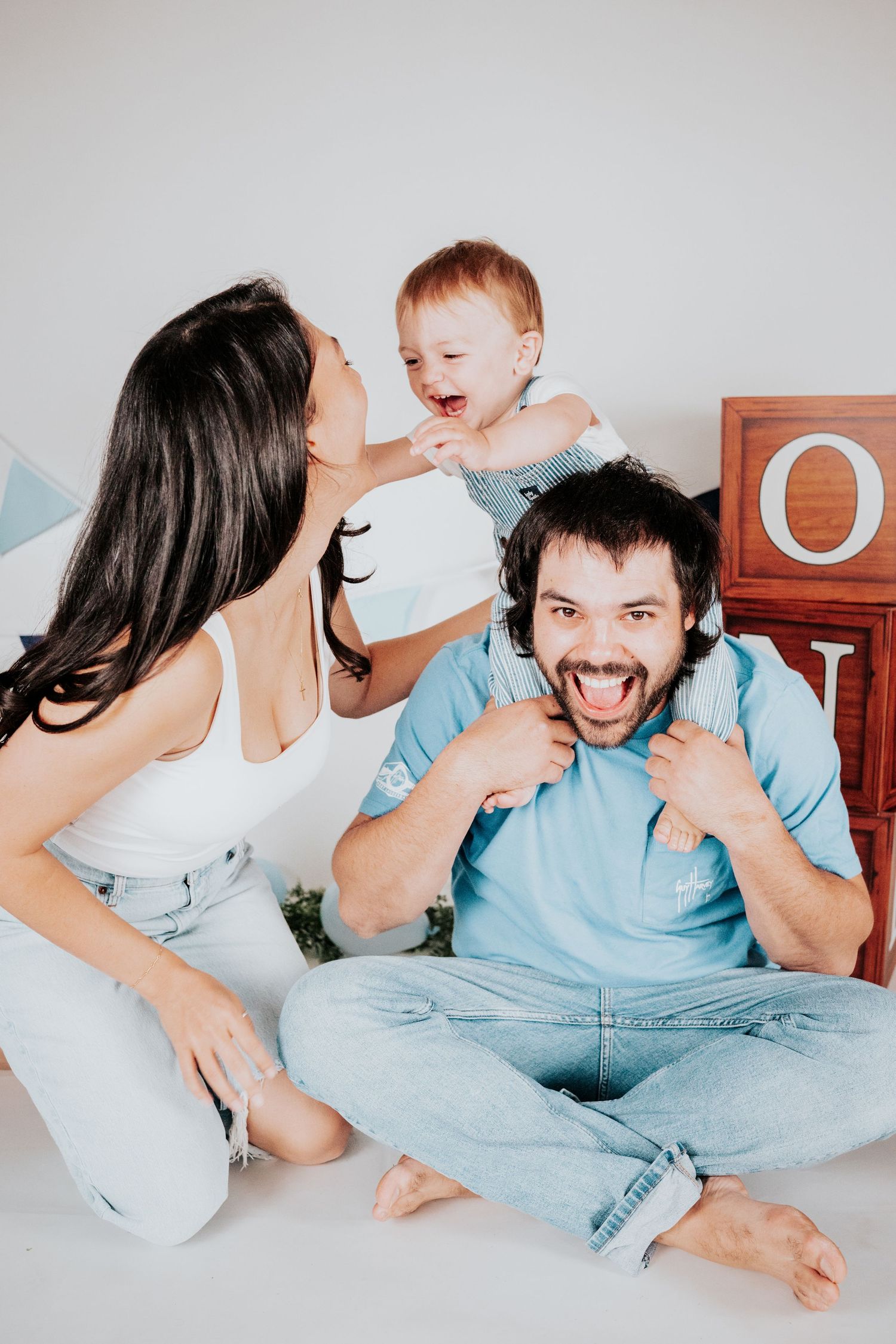 Playful family session with mom kissing baby perched on dad's shoulders, all laughing in a white photography studio.