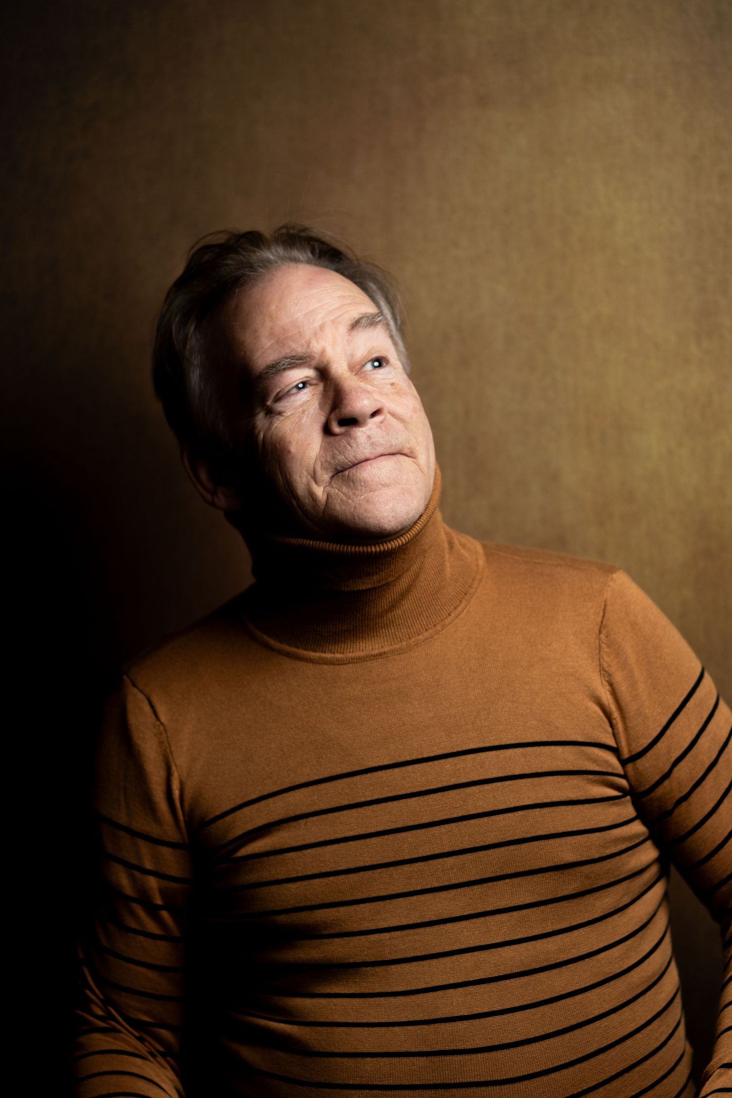 Senior man in brown striped turtleneck posing against golden background.
