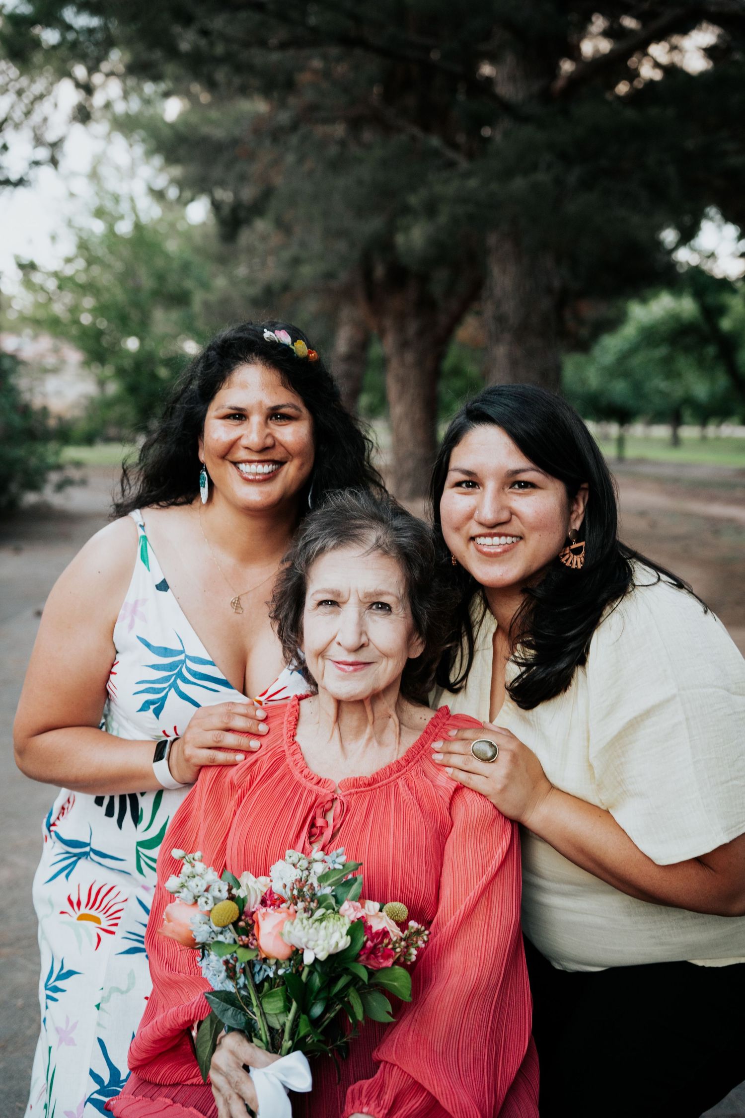 Two women smile and embrace an elderly woman holding a floral bouquet in an outdoor park setting.