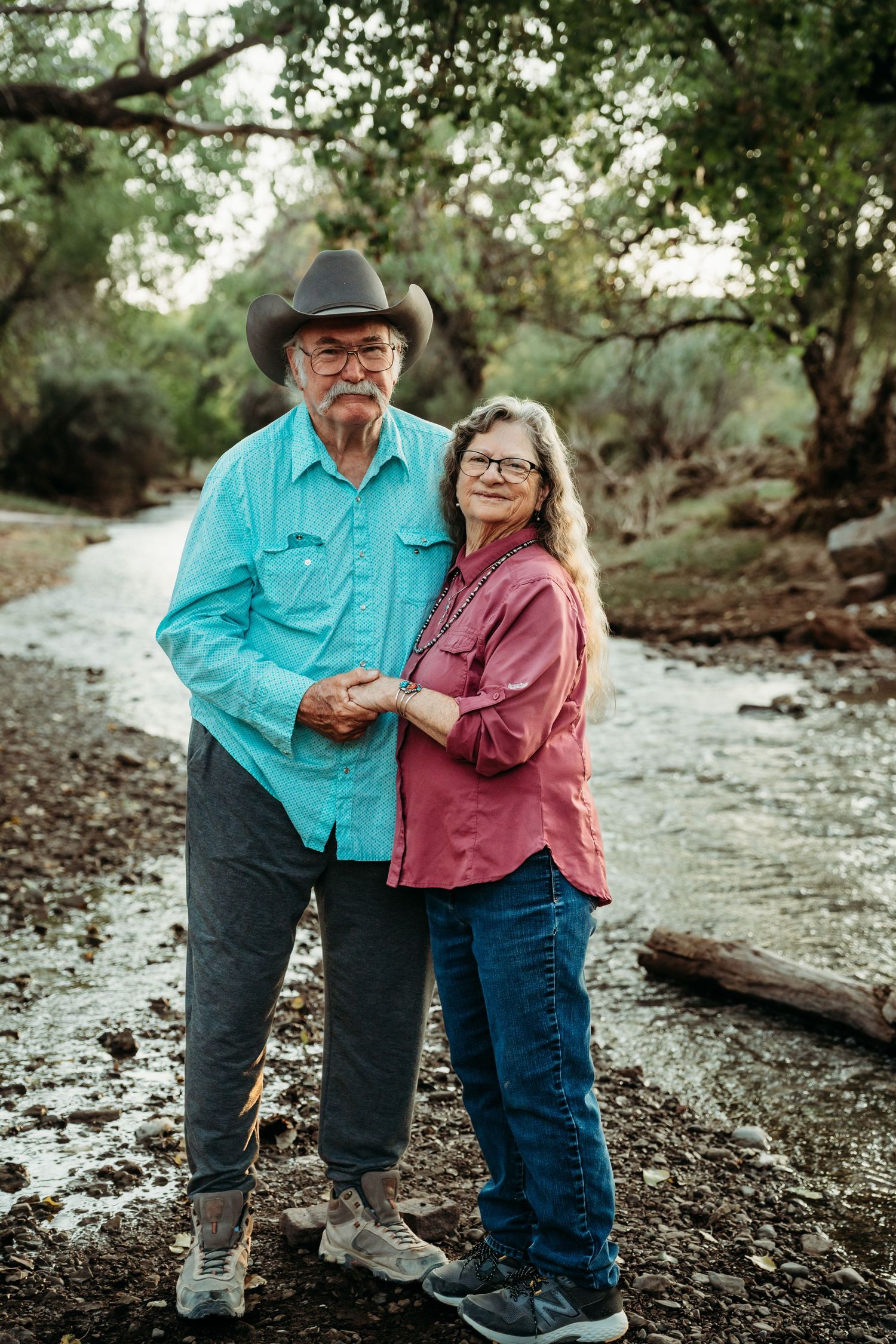 An older couple in western attire holds hands while posing near a scenic tree-lined creek.