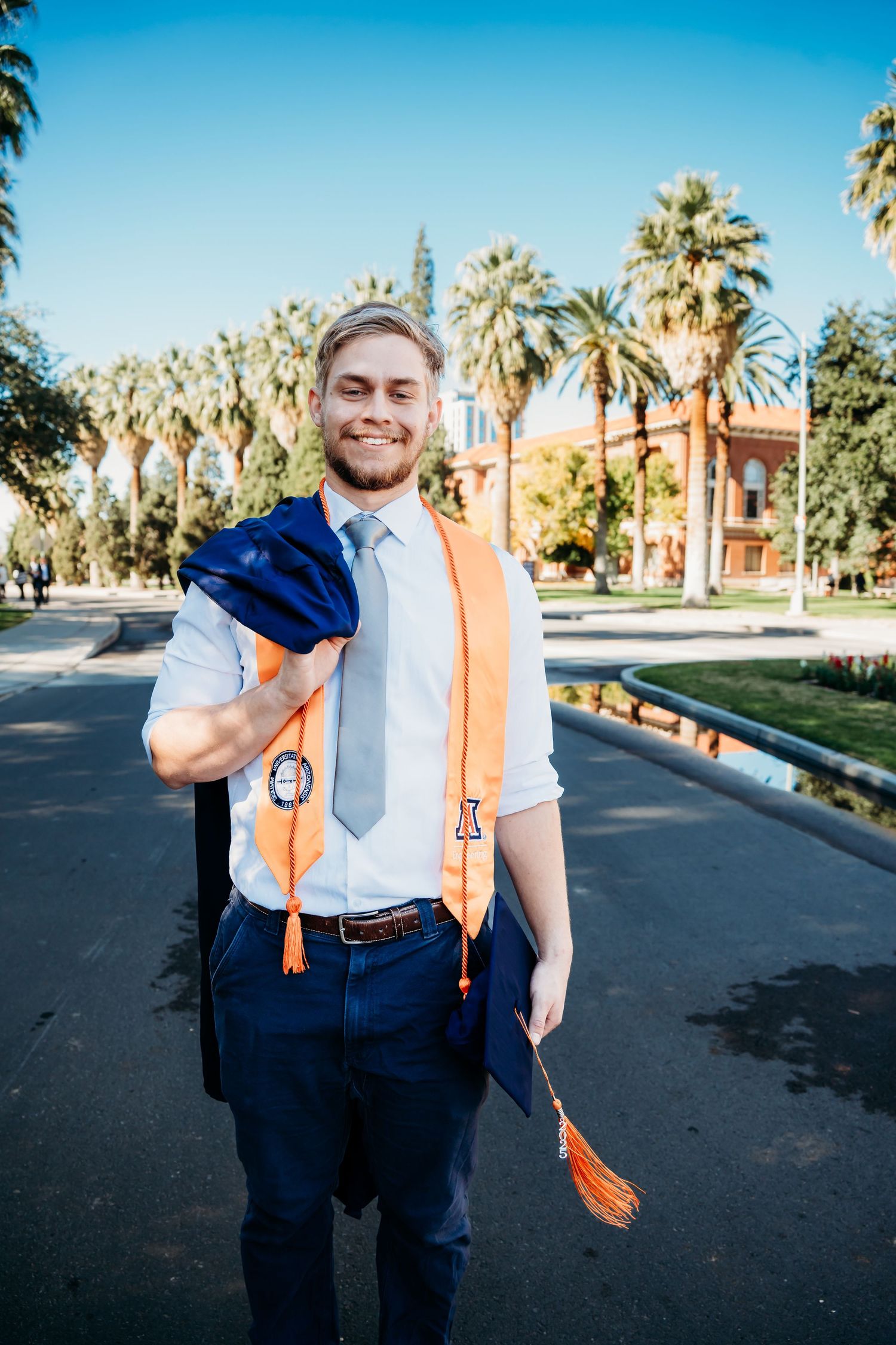 Smiling male graduate wearing orange sash holds cap outdoors near palm trees on sunny campus graduation day.