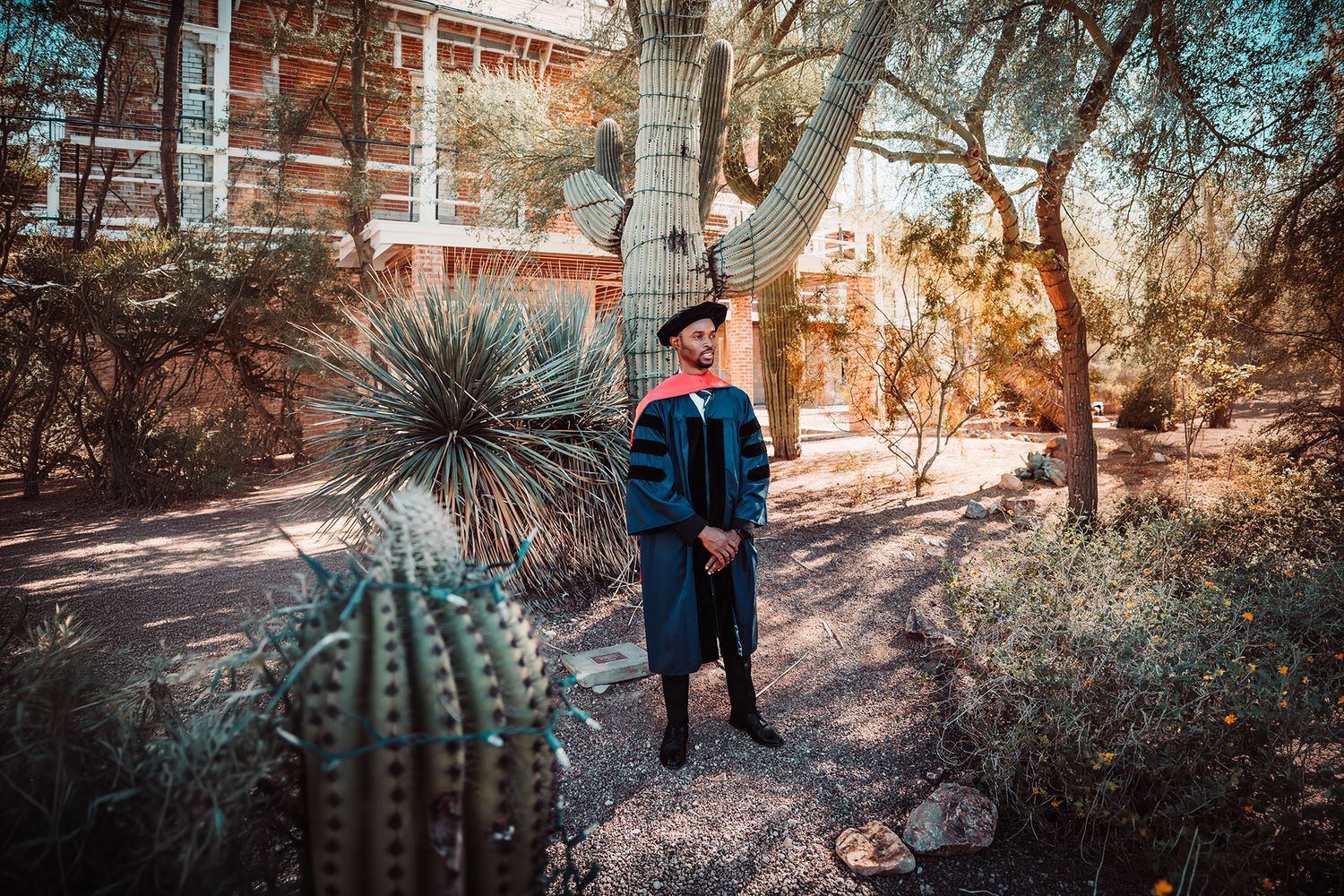Graduate in cap and gown poses among desert cacti and trees near a university building.