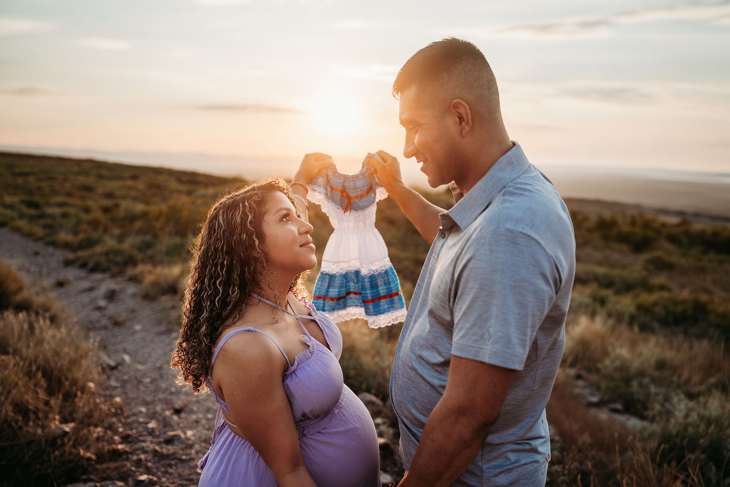Couple holding a small baby dress between them at sunset on a desert hillside during maternity shoot.