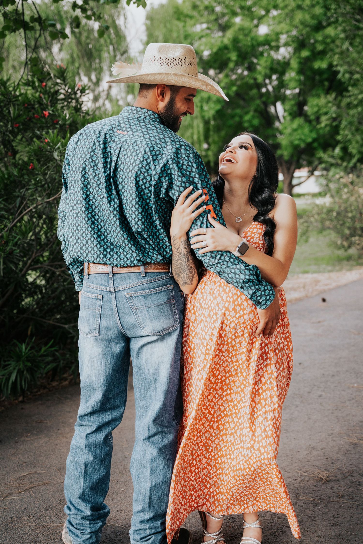 Pregnant woman in orange floral dress laughs while embracing man in cowboy hat and teal patterned shirt.