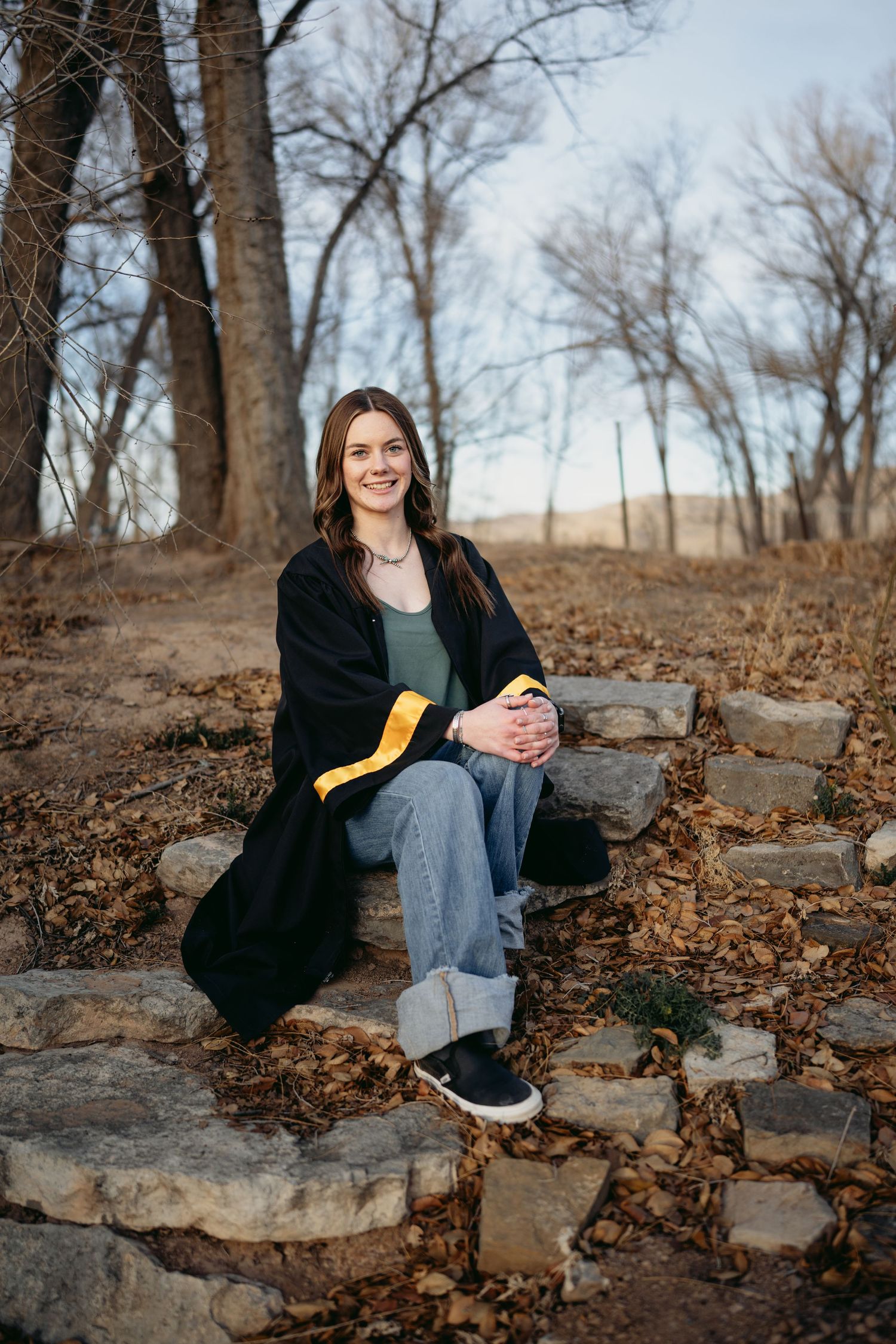 Smiling young woman in graduation gown sitting on stone steps among autumn fallen leaves.