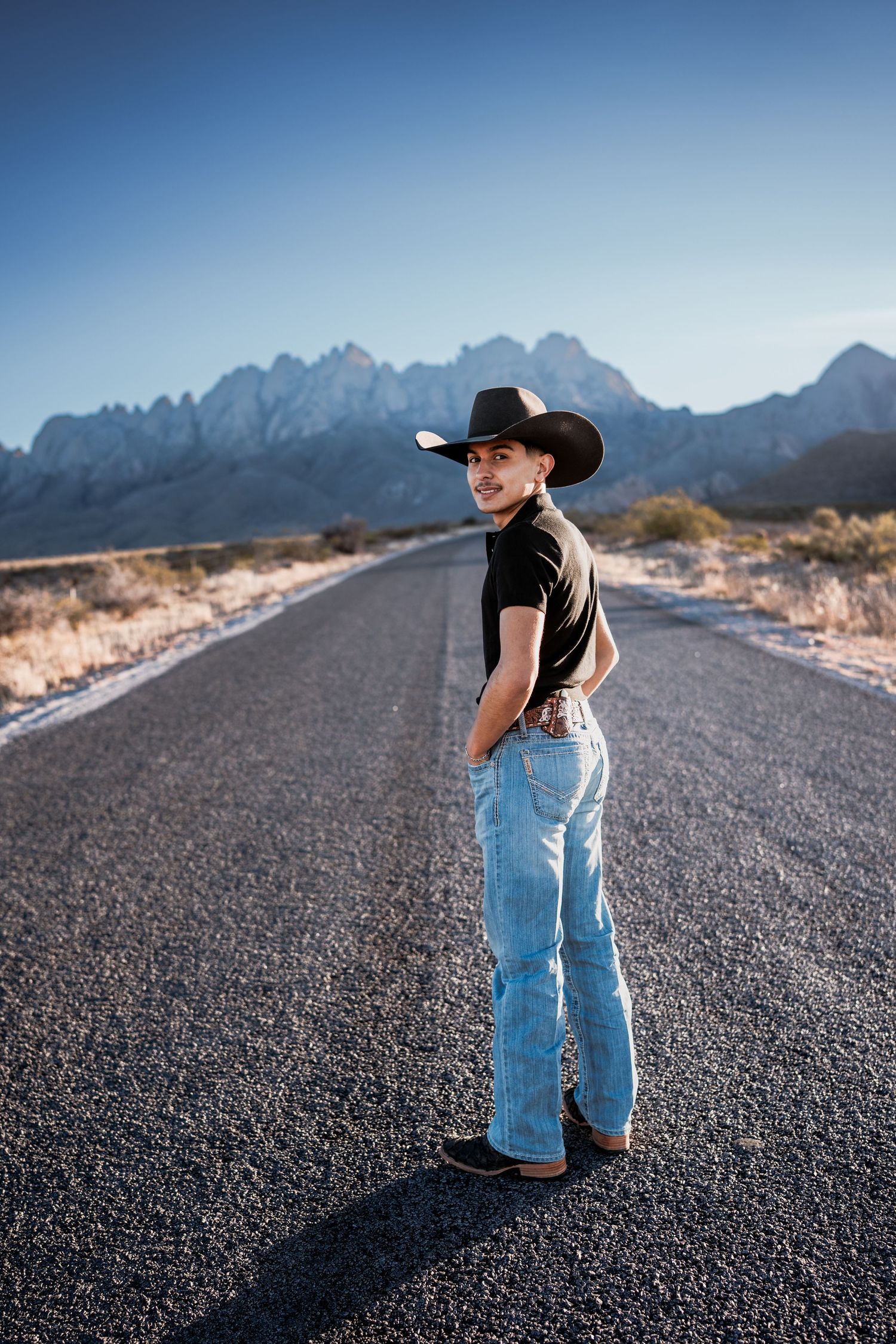 Young man in black cowboy hat and jeans standing on open desert road with mountains behind.