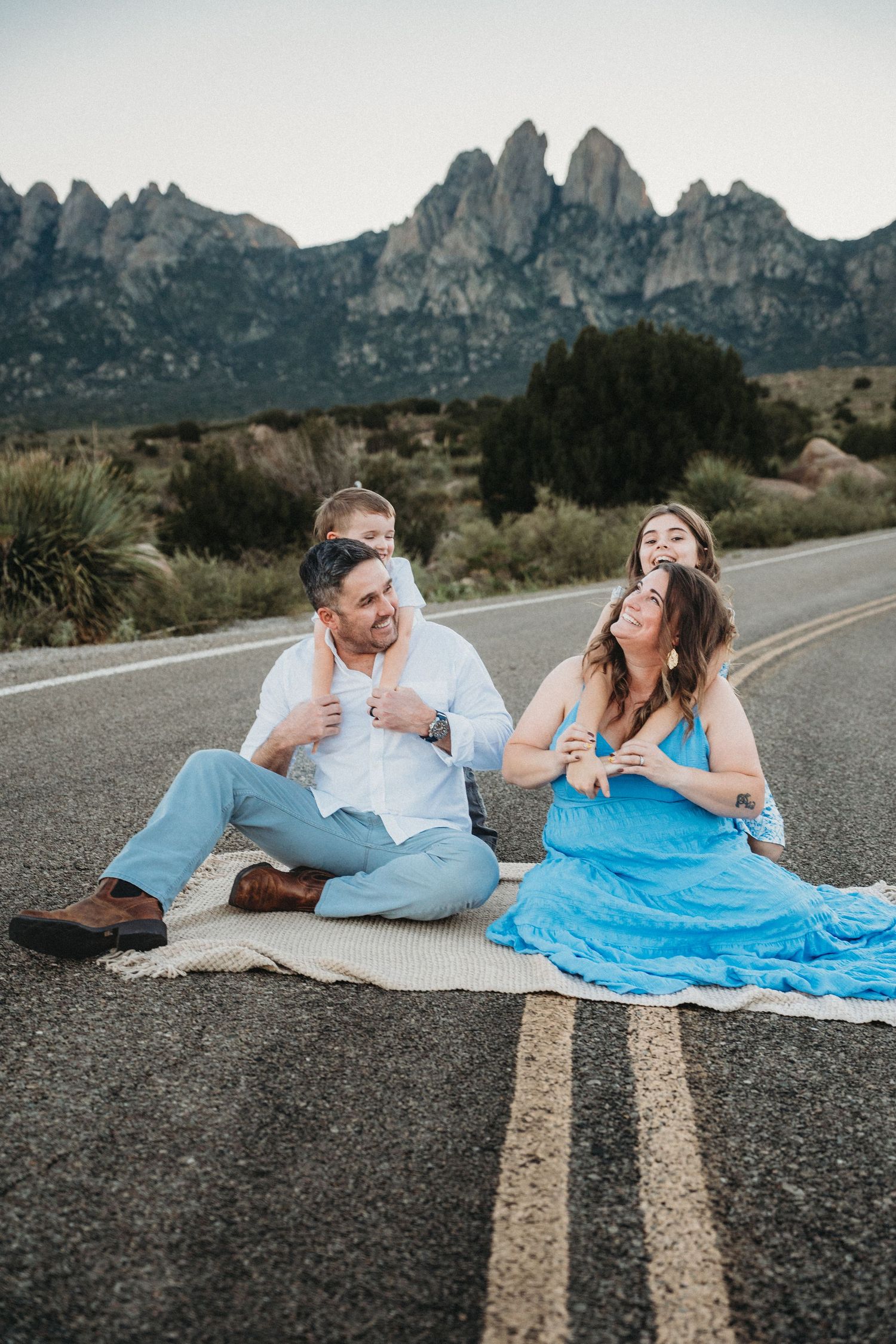 Happy family sitting on a blanket on a road with rocky mountains and desert landscape behind them.