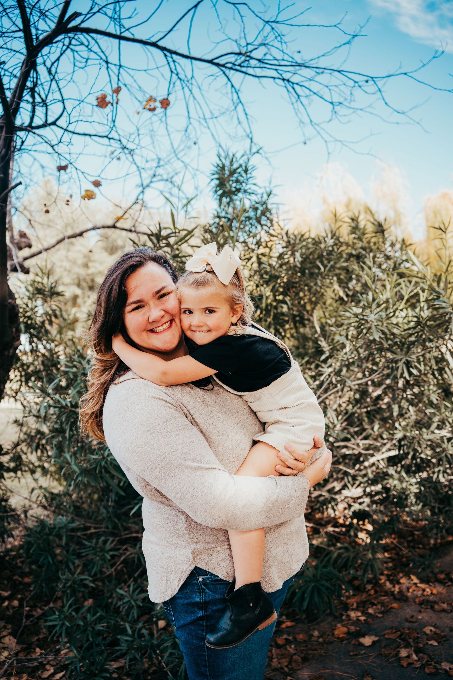 Smiling mother holding toddler girl with bow in hair, embracing outdoors near green shrubs and trees.