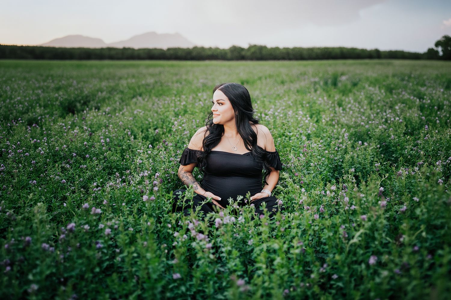 Pregnant woman in black off-shoulder dress sitting in blooming green field at sunset.