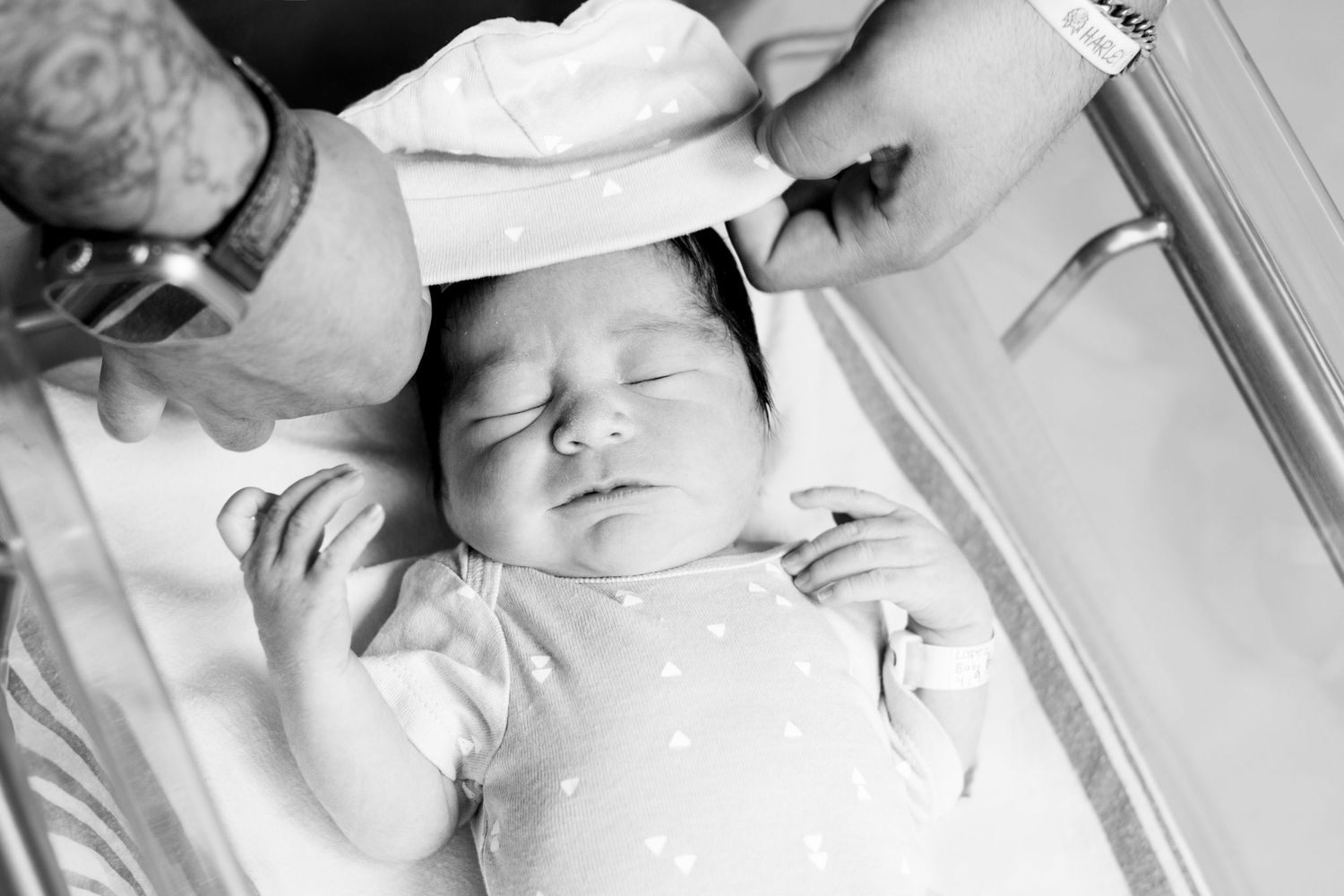 Black and white photo of a newborn baby in a hospital bassinet having a hat placed on head.