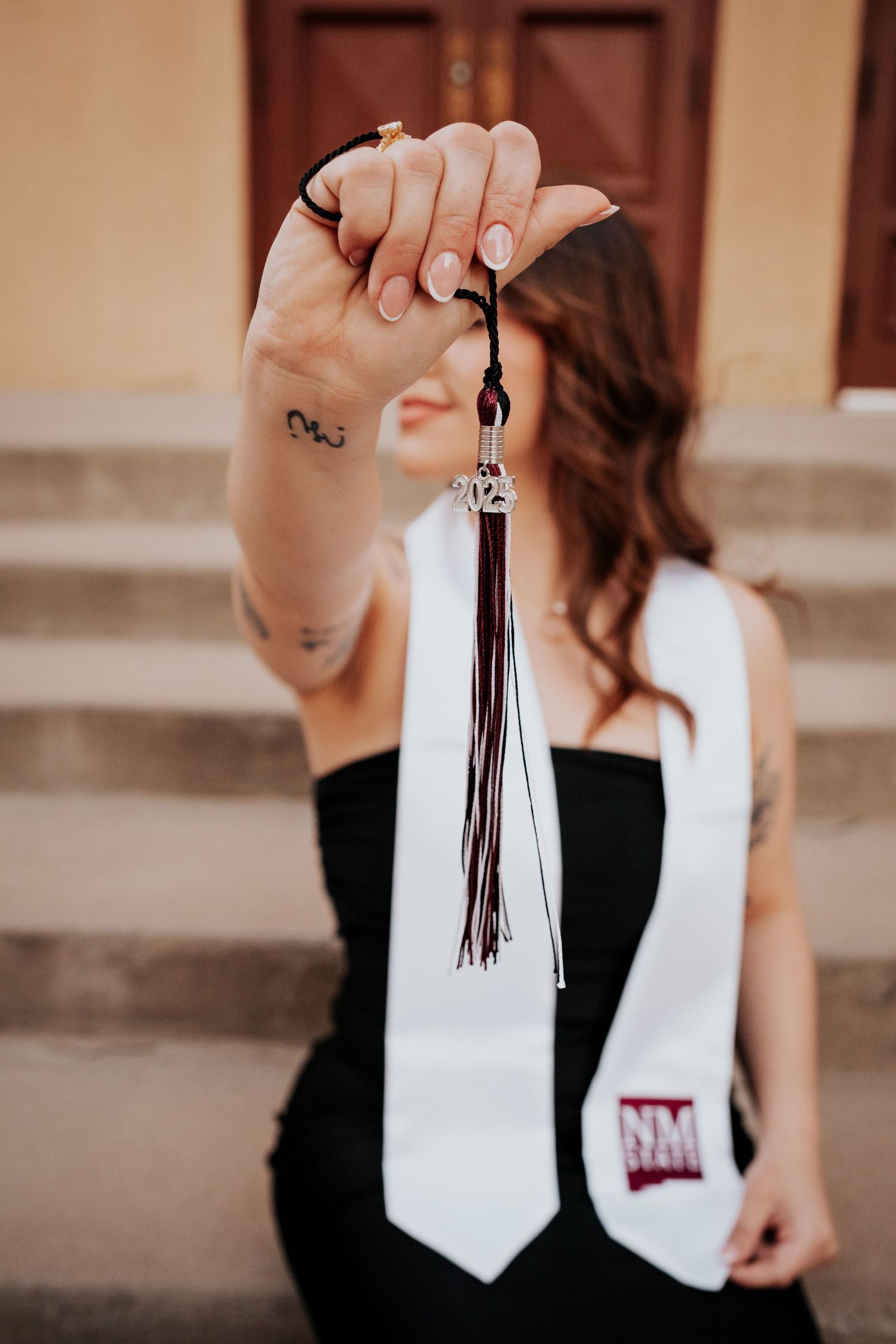 Graduate holding maroon tassel toward camera with white NM State stole visible in background.