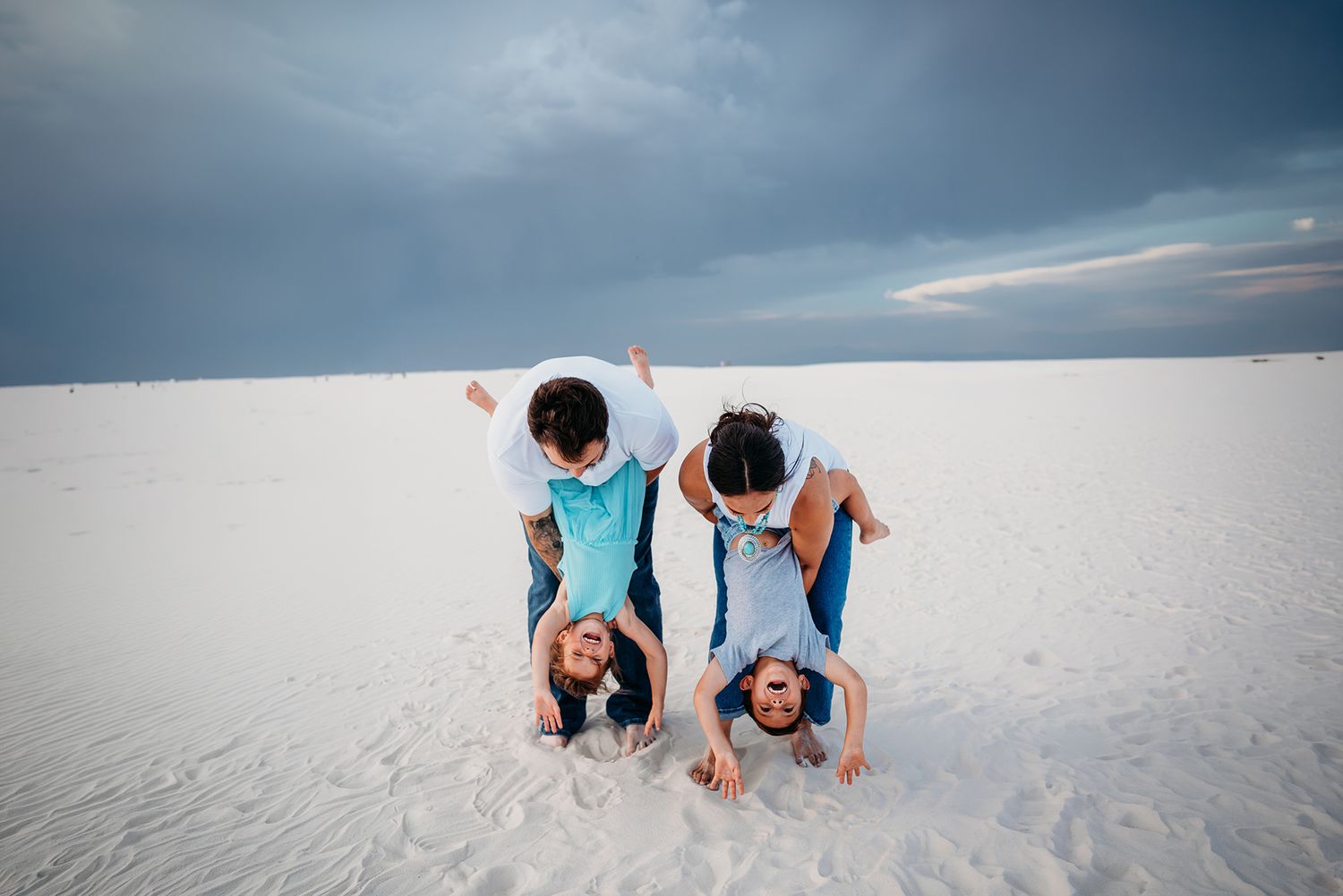 Two adults playfully dip two children upside down on white sand dunes under a cloudy sky.