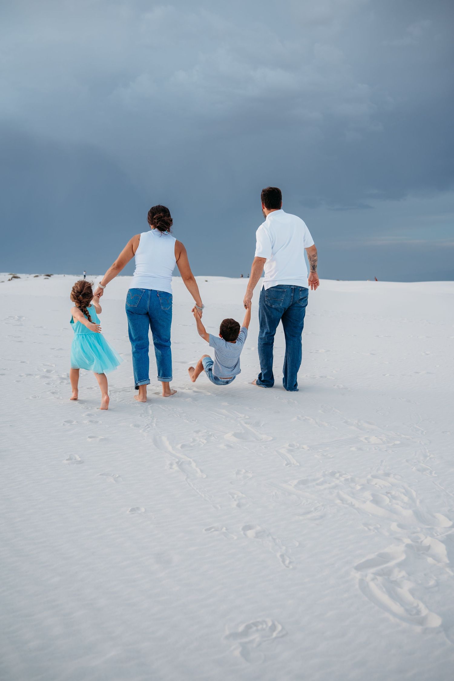 A family of four walks hand-in-hand across white sand dunes under a dramatic cloudy sky.