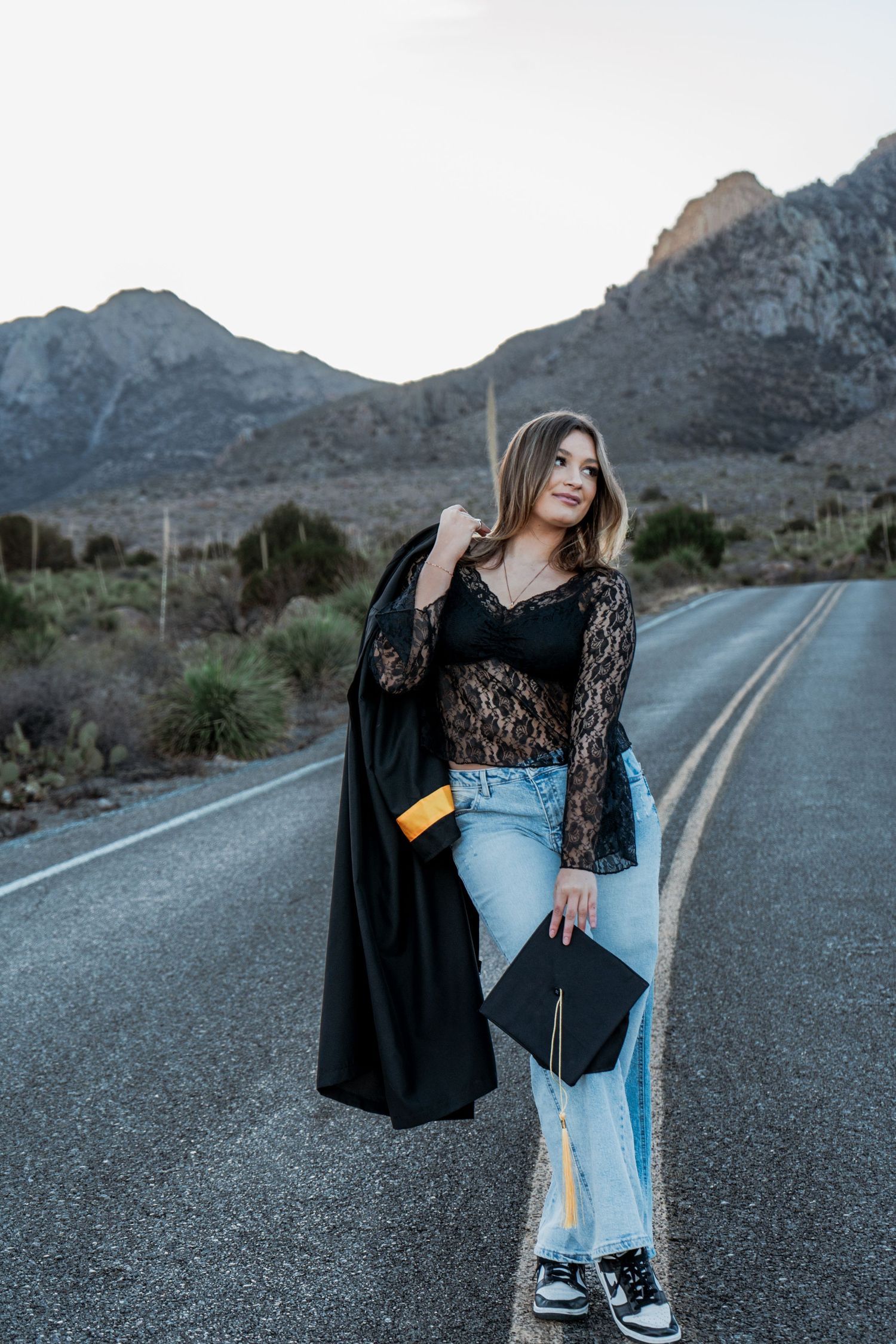 Graduate holds cap and drapes gown over shoulder while posing on mountain road at sunset.