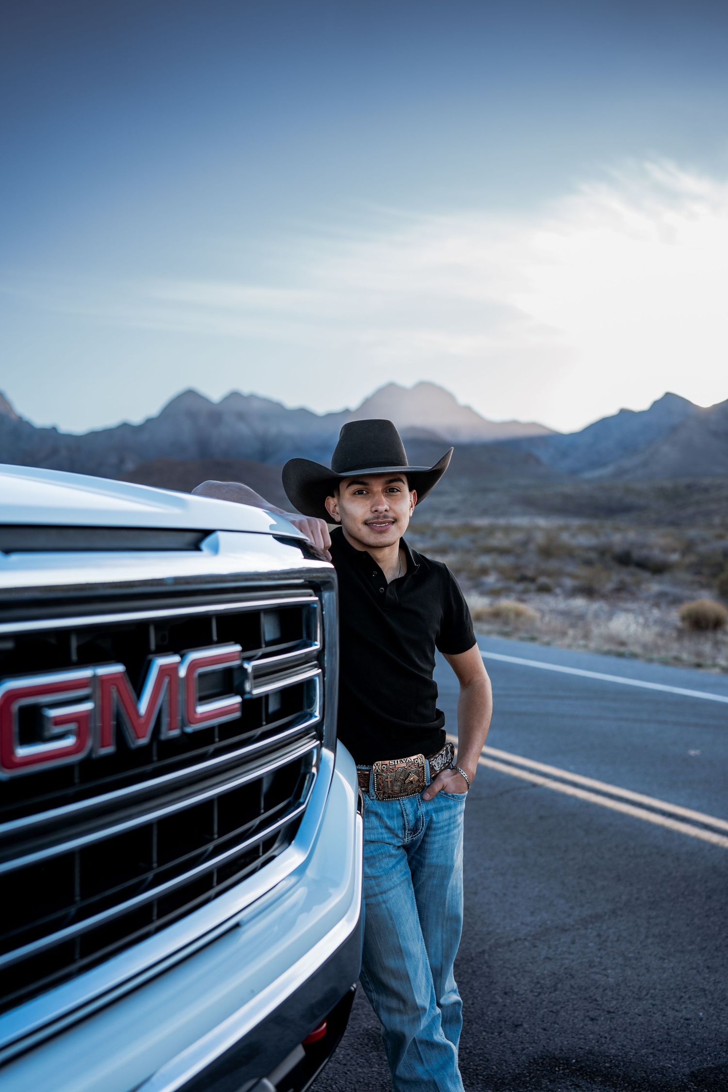 Young man in cowboy hat leaning against white GMC truck grille on desert road.