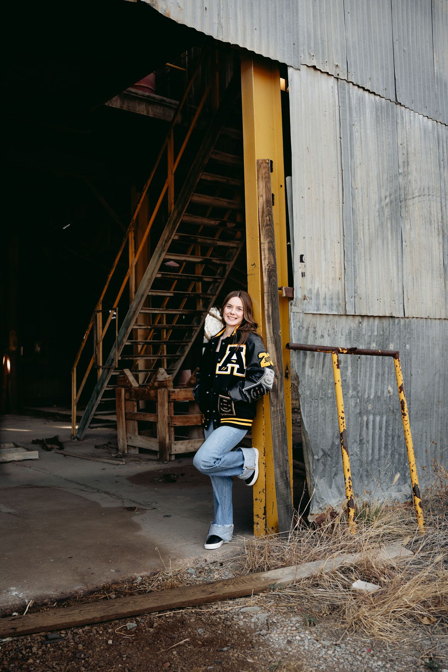 High school senior girl in black varsity jacket posing in doorway of rustic industrial building.