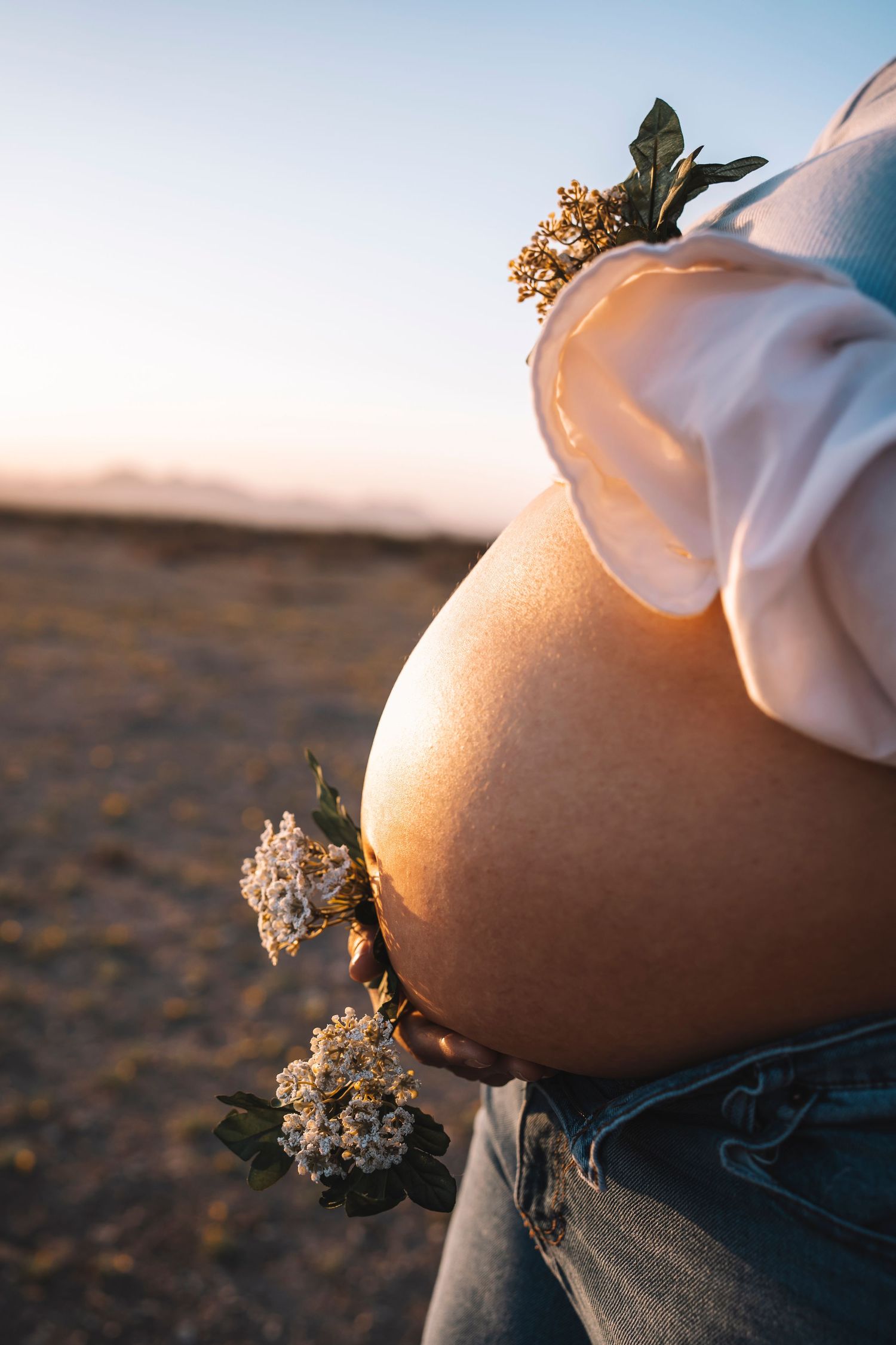 Close-up of a pregnant woman's bare baby bump adorned with small white wildflowers against a warm sunset sky.