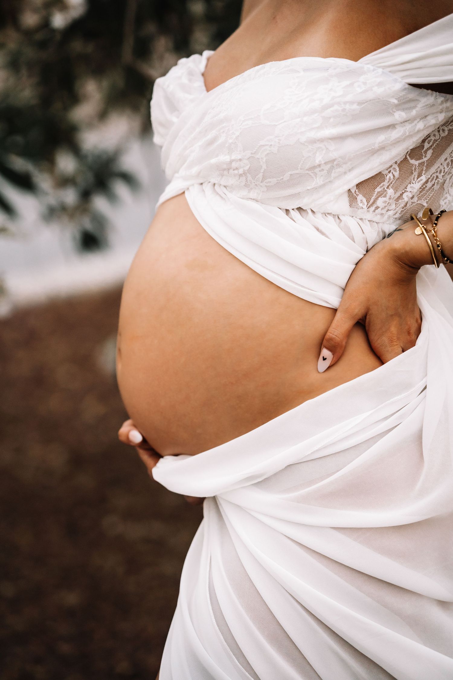 Close-up of pregnant woman in white off-shoulder dress with gold bracelets cradling her bare baby bump.