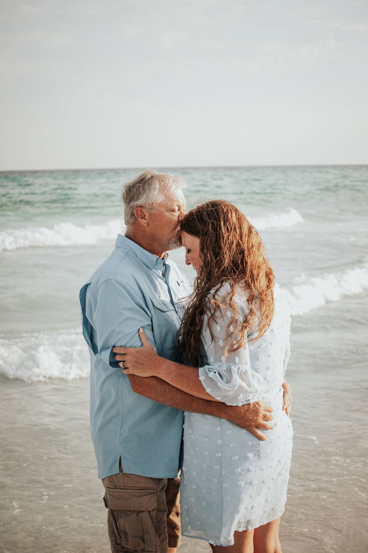 Older couple sharing a tender forehead kiss while embracing on a beach with turquoise ocean waves.