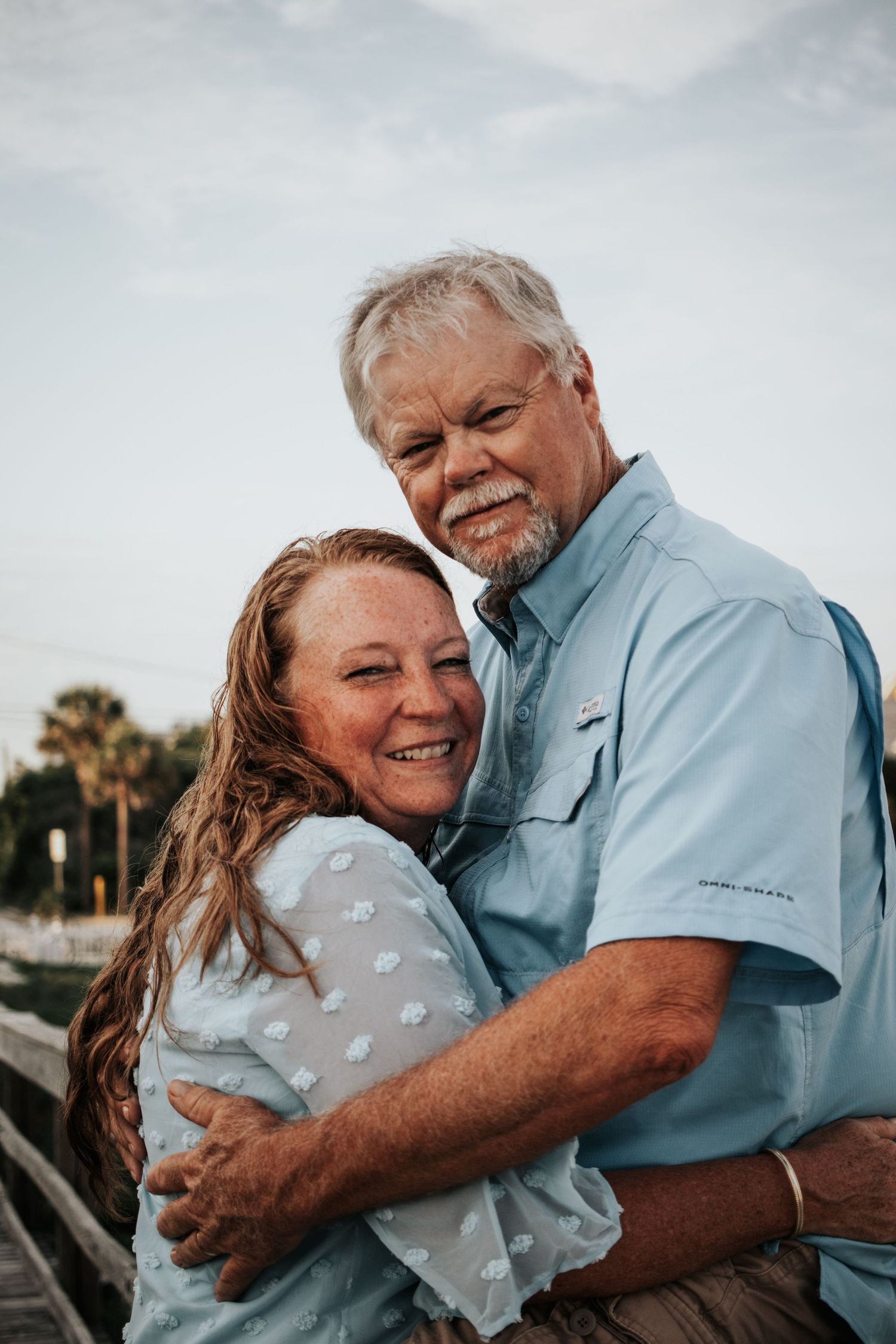 Happy mature couple hugging and smiling at camera on wooden pier, man in light blue fishing shirt.