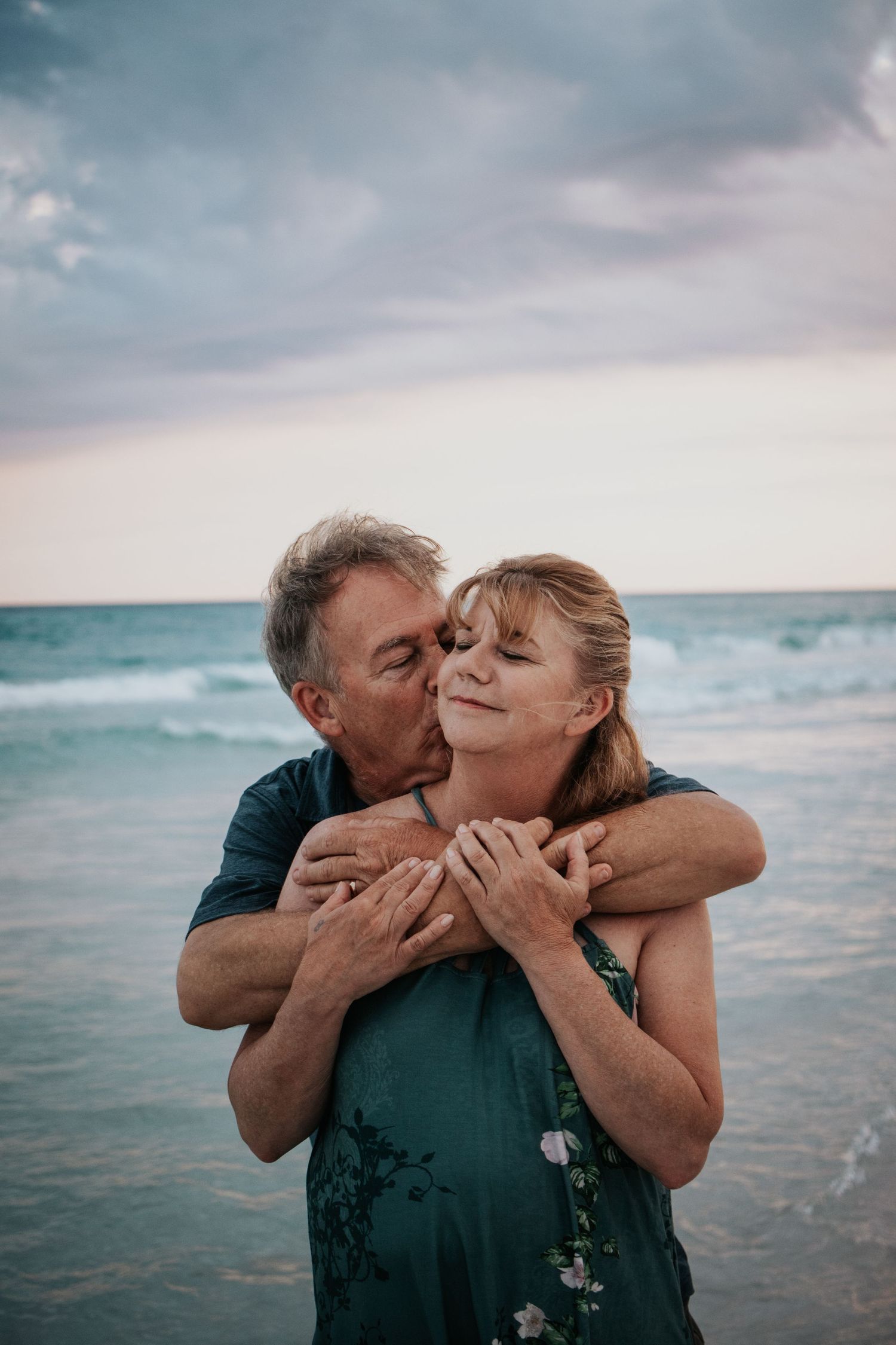 Man kissing woman on the cheek while embracing her from behind on a beach at sunset.