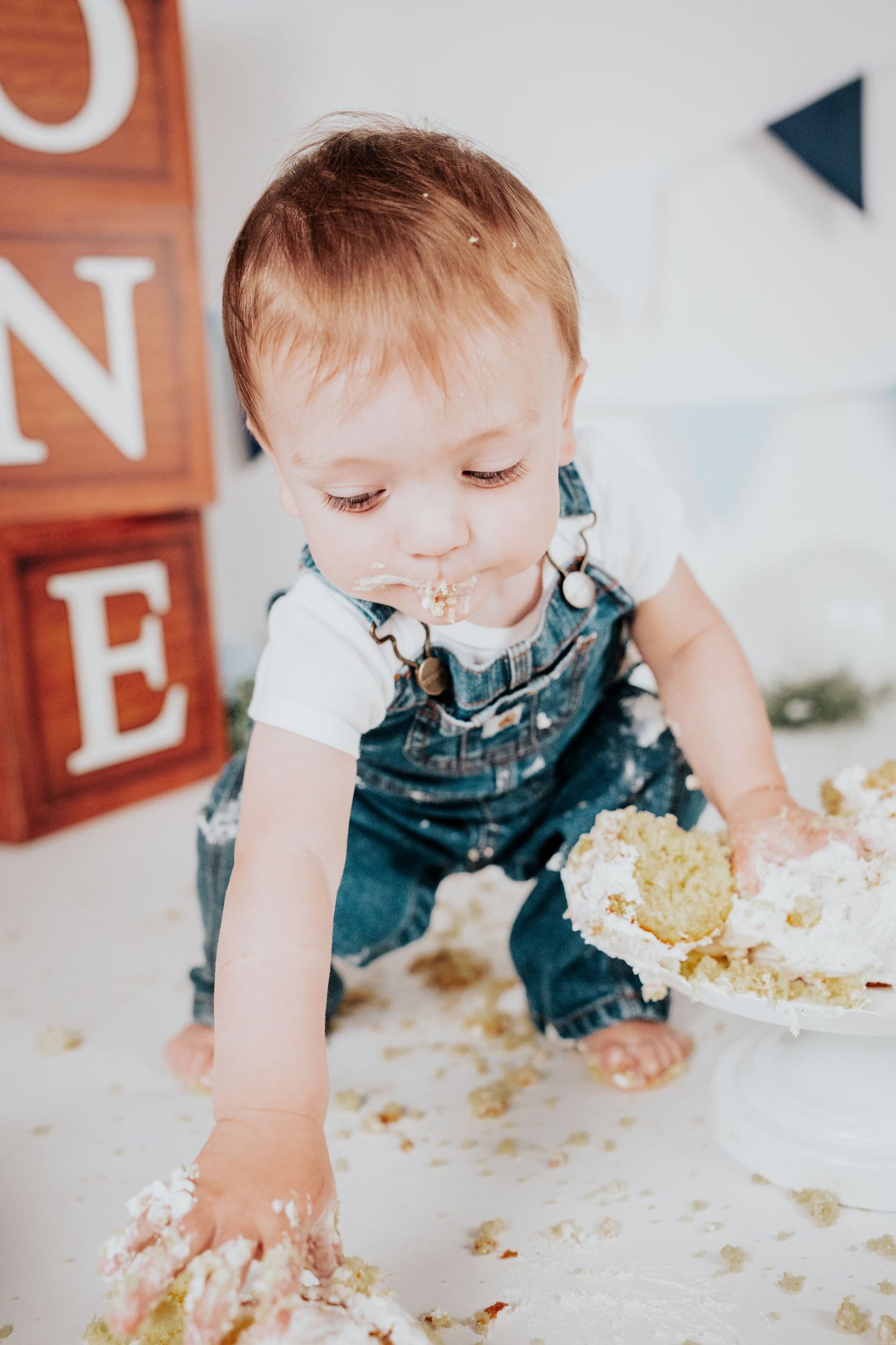 Messy-faced baby in Carhartt overalls reaching into smashed first birthday cake on white floor.