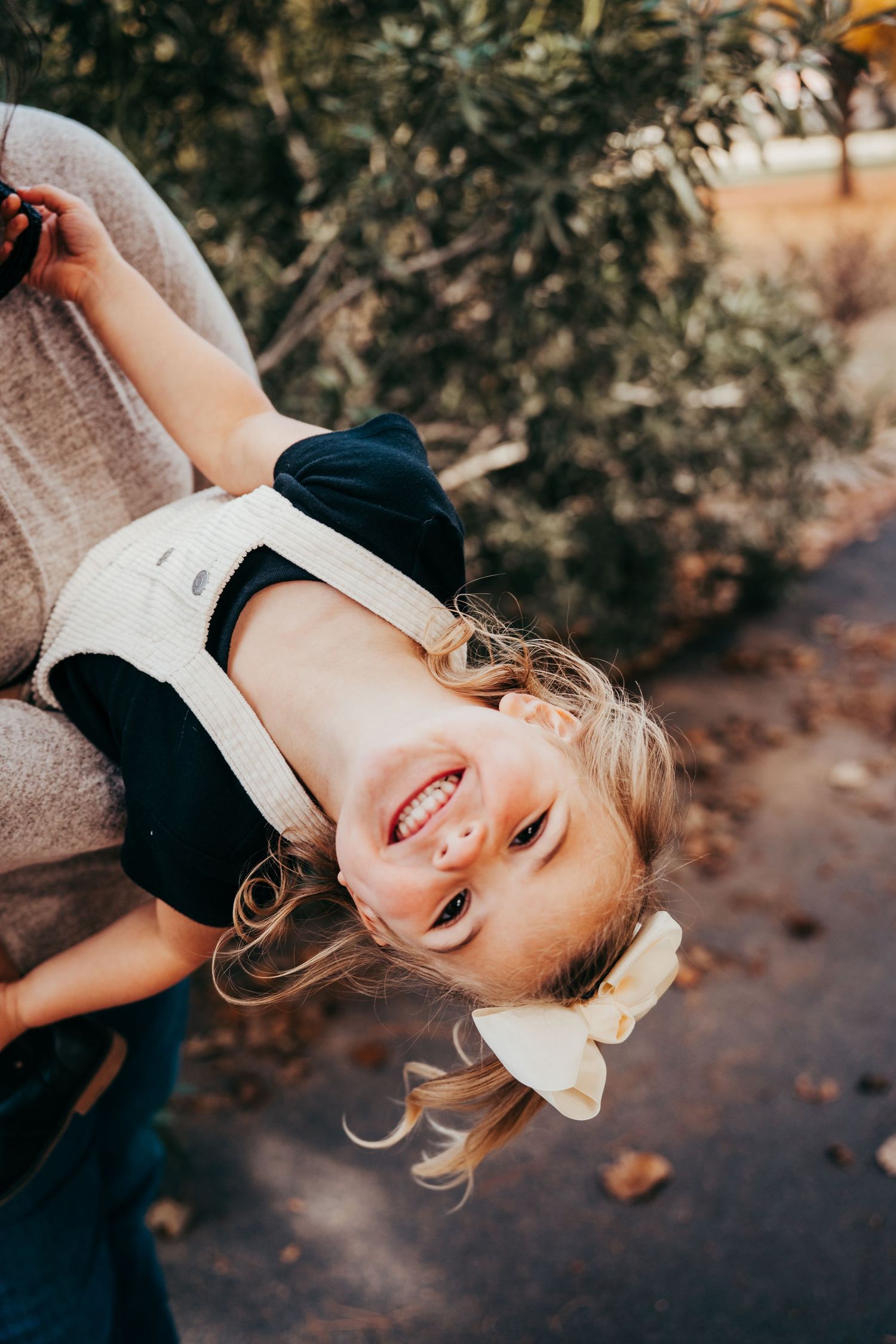 Joyful young girl with blonde hair and bow laughing upside down while being held by her mother.