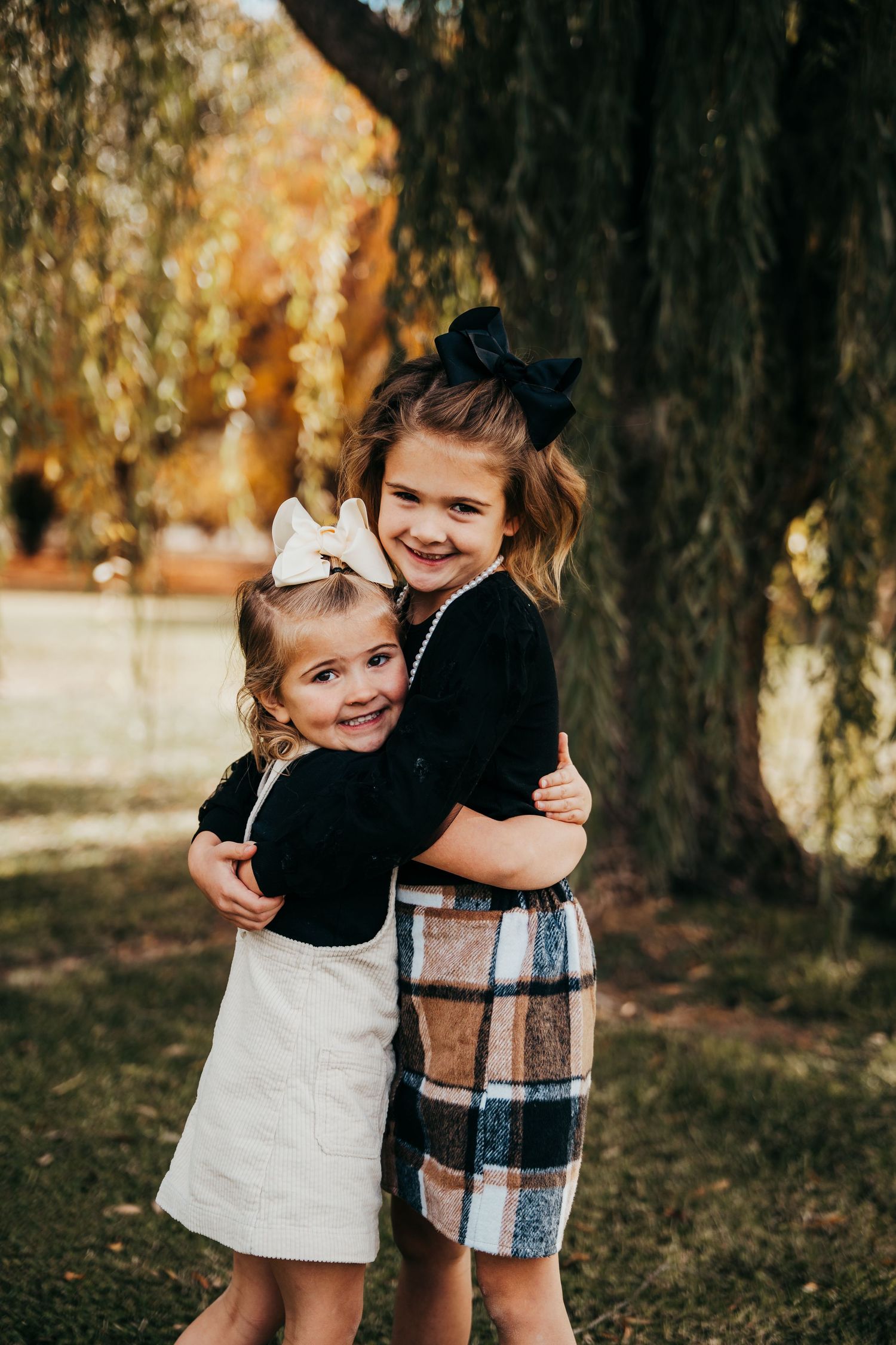 Two young girls in black outfits with hair bows share a warm hug outdoors beneath a willow tree in autumn.