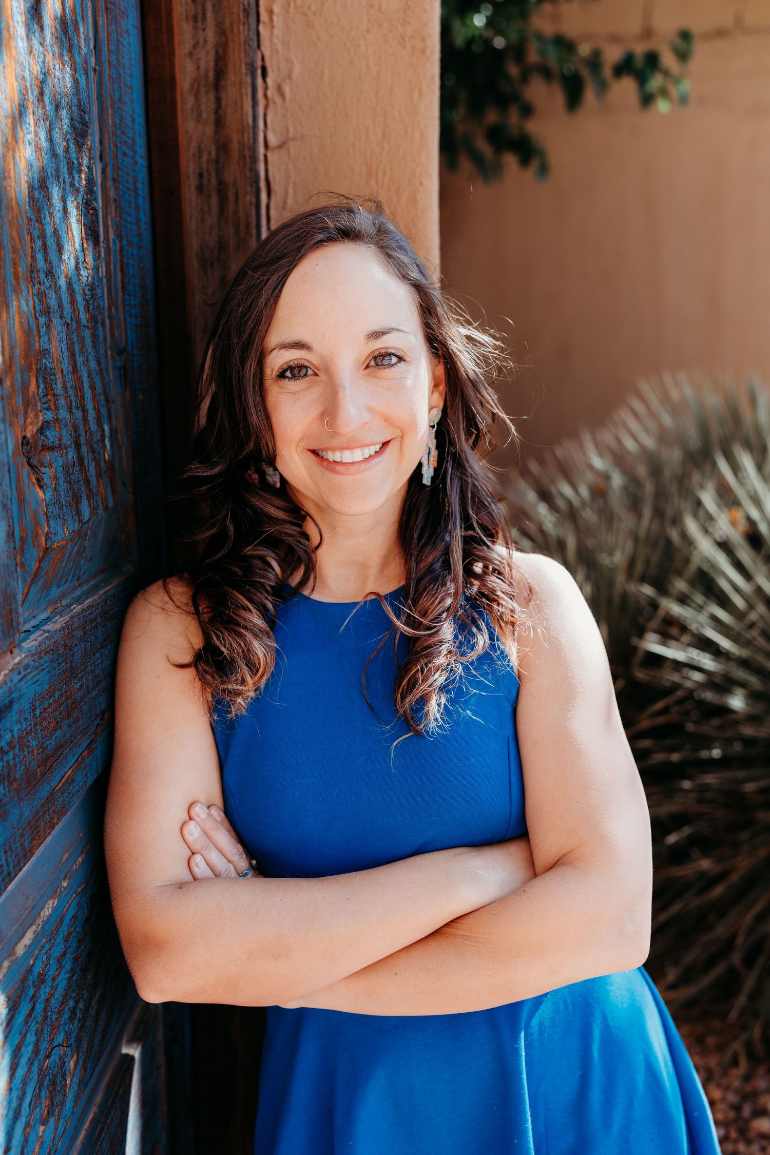 A smiling woman with dark hair in a blue dress leans against a rustic wooden door with arms crossed.