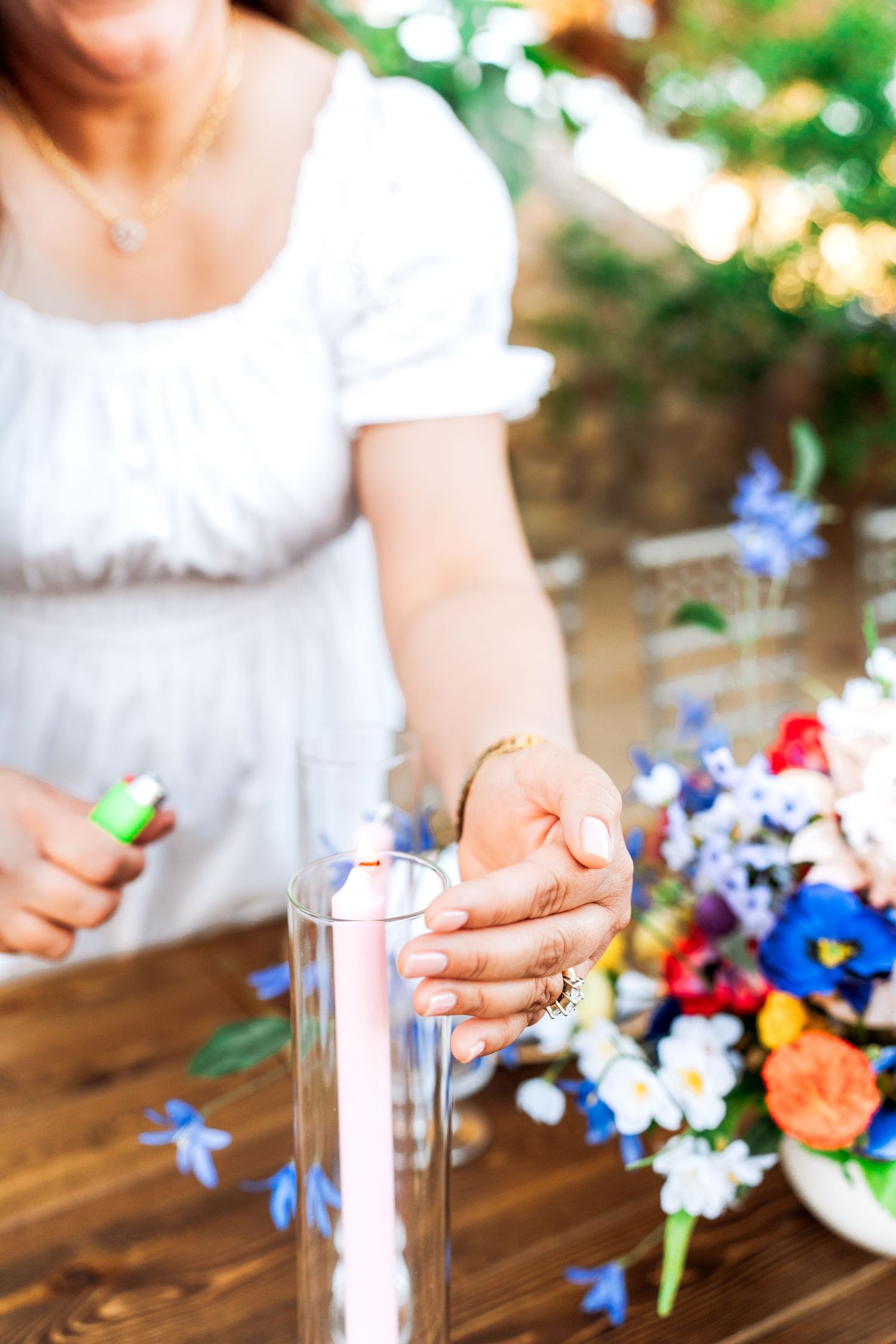 Woman in white dress lighting a pink taper candle in a glass vase beside colorful wildflower centerpiece at wedding table.