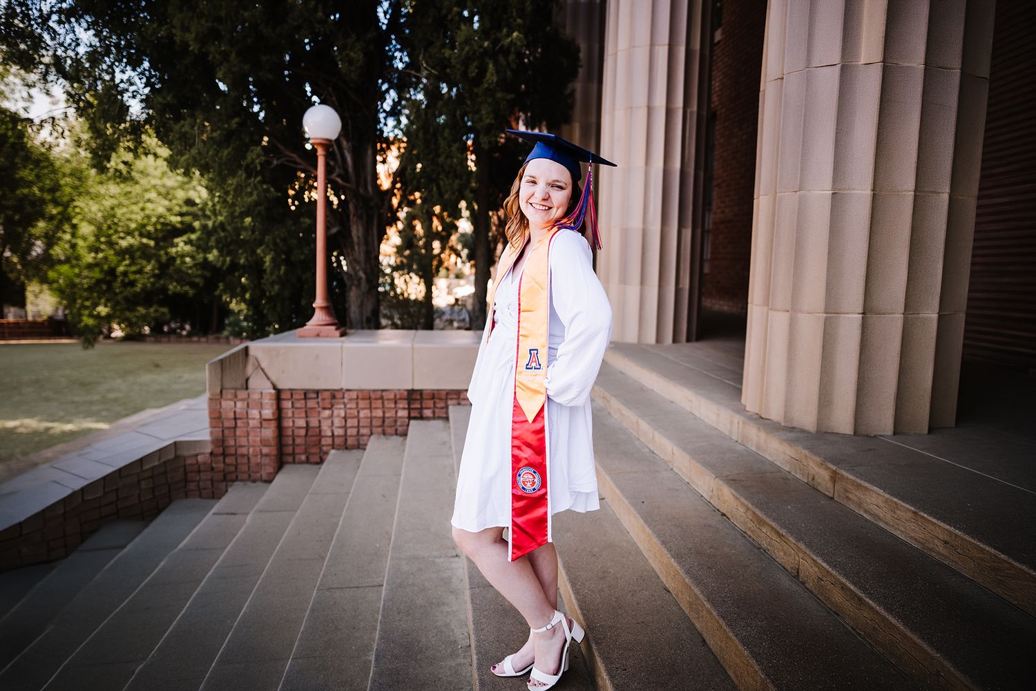 Young woman in white graduation gown and blue cap poses on university steps with red sash.