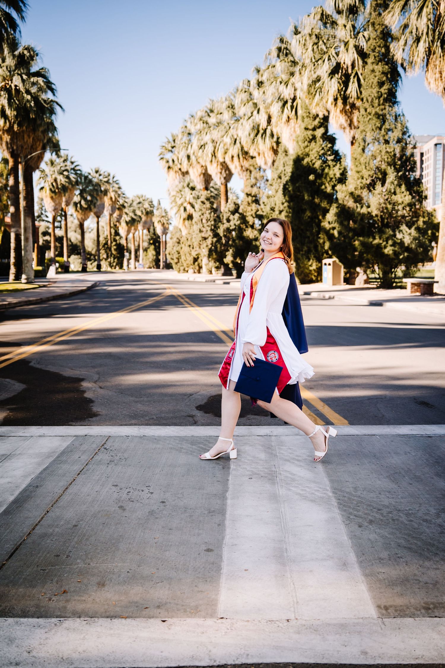 Young woman in graduation gown crossing a palm-lined street, smiling confidently in heels on a sunny day.