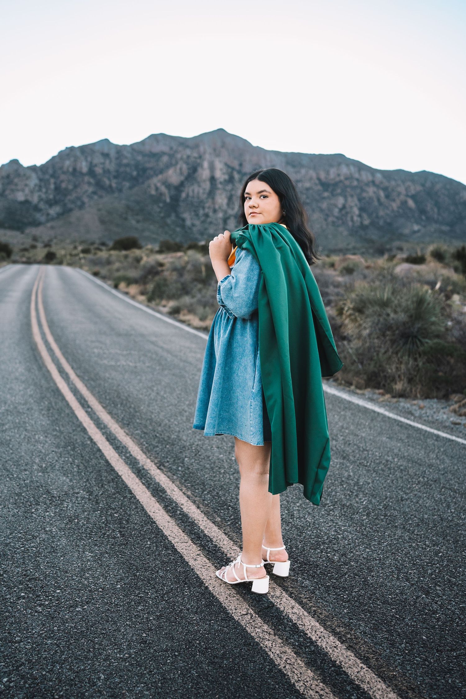 Young woman in a denim dress holding a green graduation gown on a desert highway with mountain backdrop.