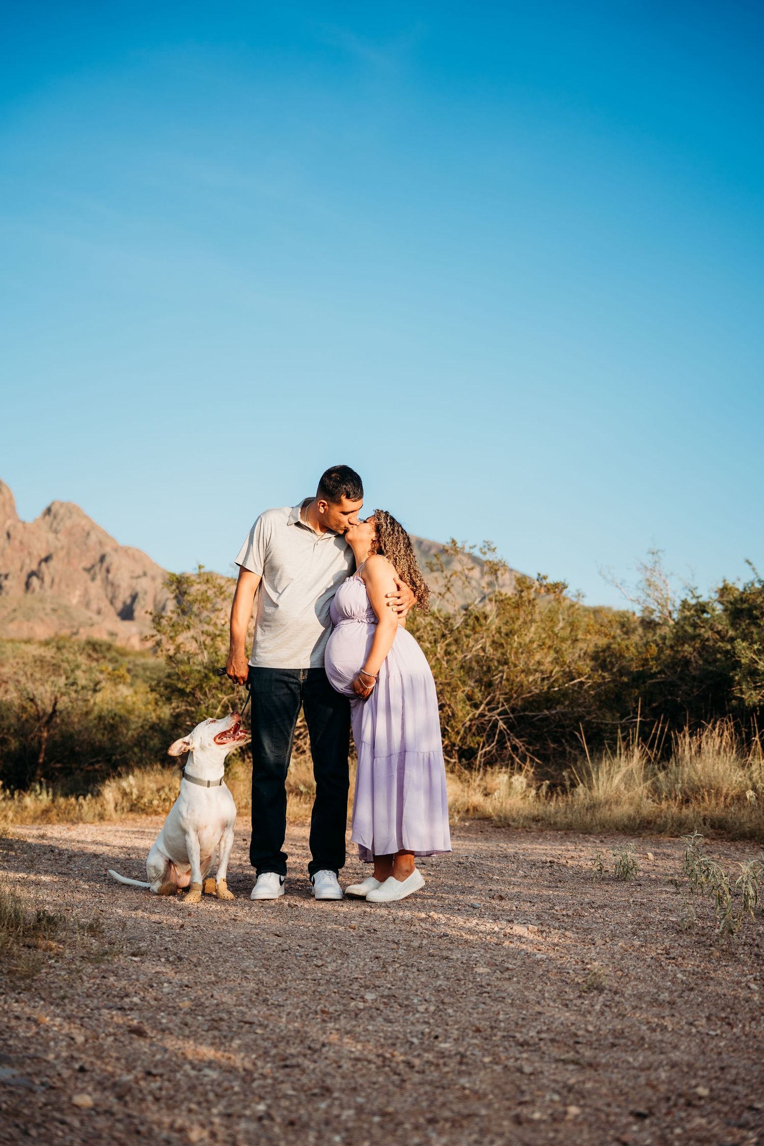 Expecting couple kissing on desert trail with white dog beside them, rocky mountains and blue sky backdrop.
