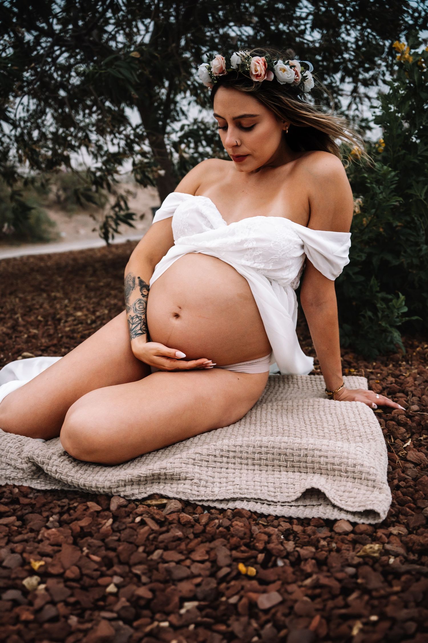 Pregnant woman wearing floral crown and white off-shoulder top sits on blanket tenderly holding her bump.